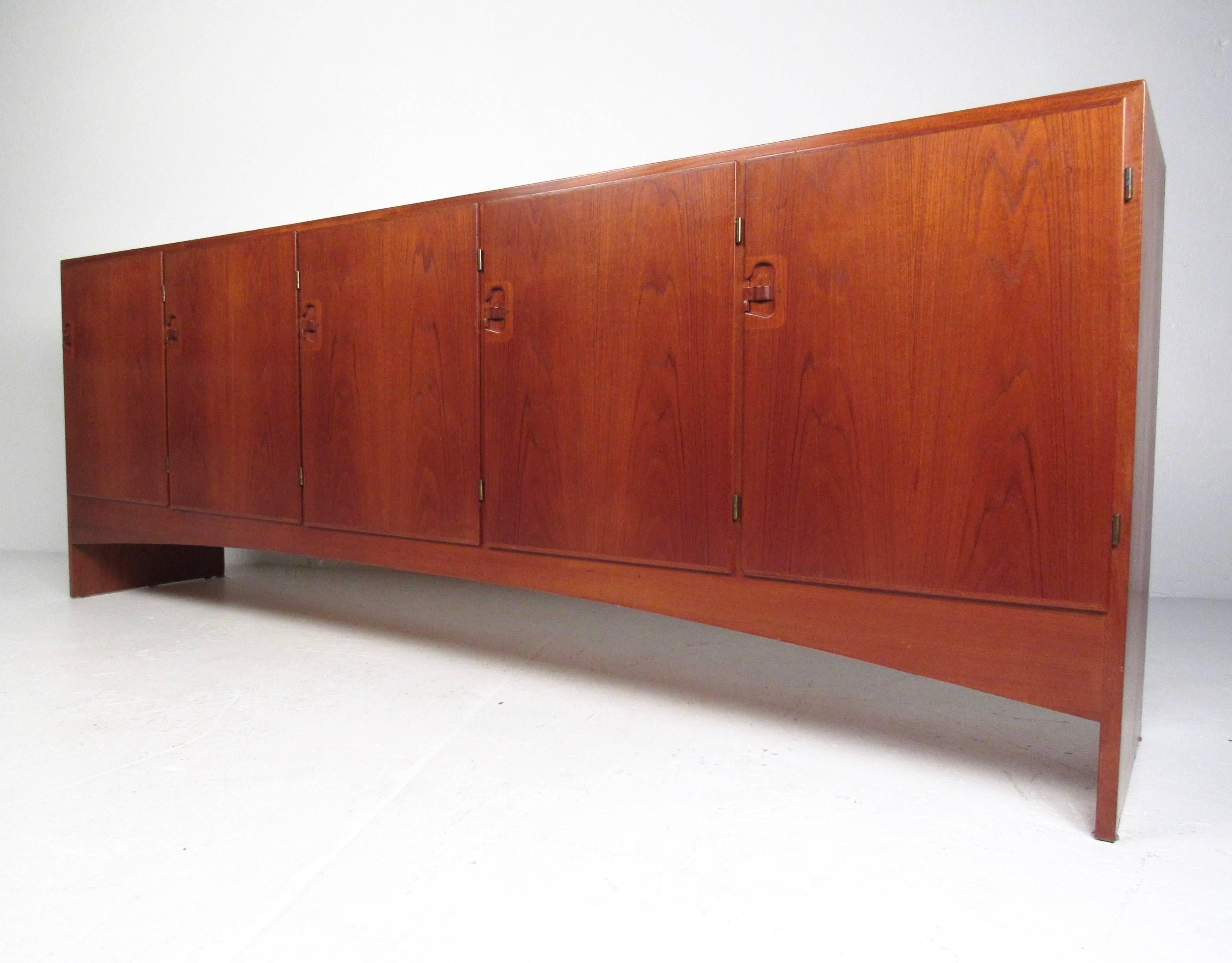 This long sideboard features a beautiful vintage teak finish with five spacious cabinets for storage in any setting. Ideal mid-century credenza for office use or dining room storage. Adjustable shelves, dovetailed drawers, and unique drop-latch door