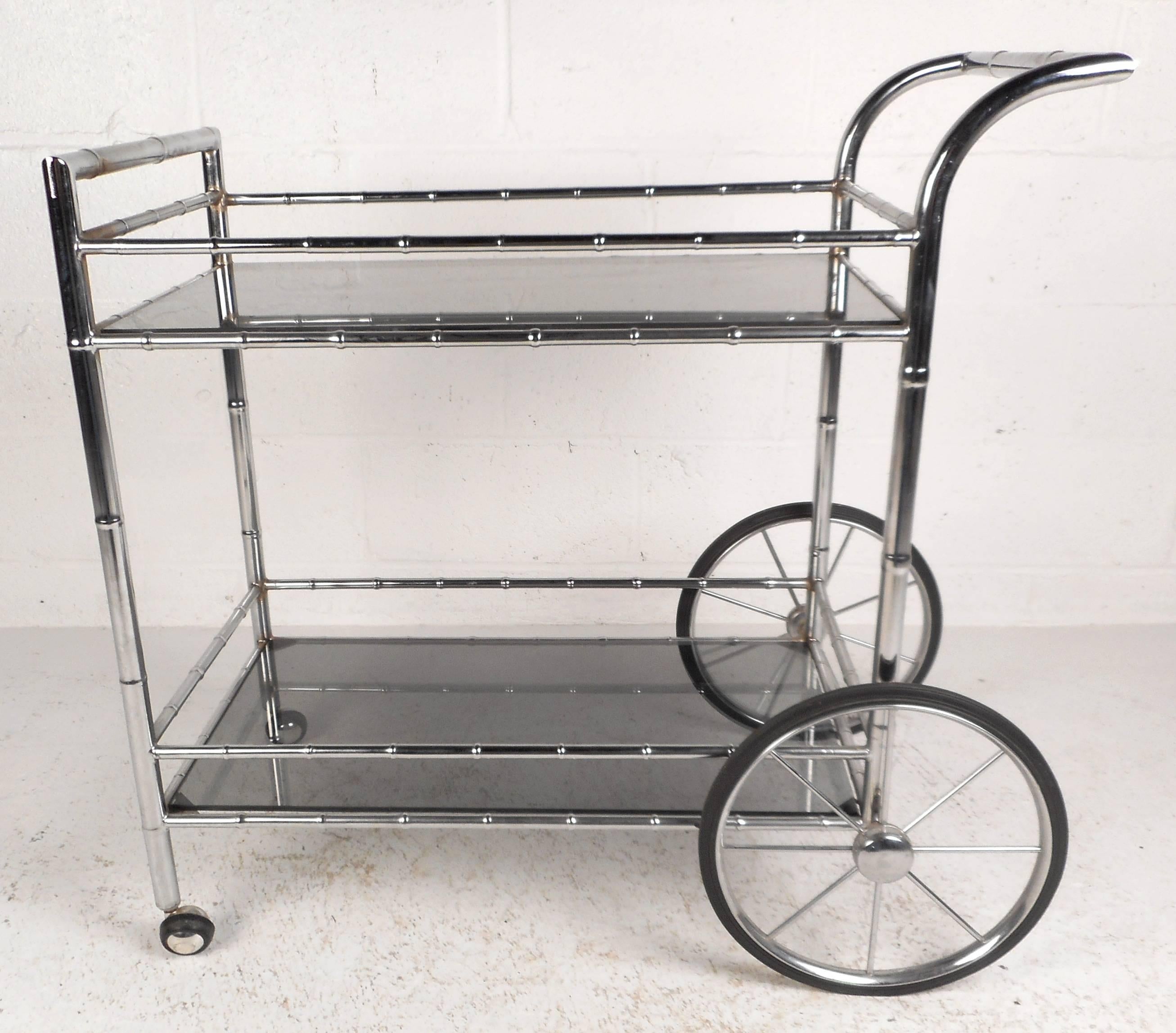 Elegante carrello da bar in stile vintage moderno con struttura in finto bambù cromato, grandi ruote a raggi e ripiani in vetro fumé. Il magnifico design a due livelli è dotato di ulteriori barelle per evitare che gli oggetti cadano mentre sono in