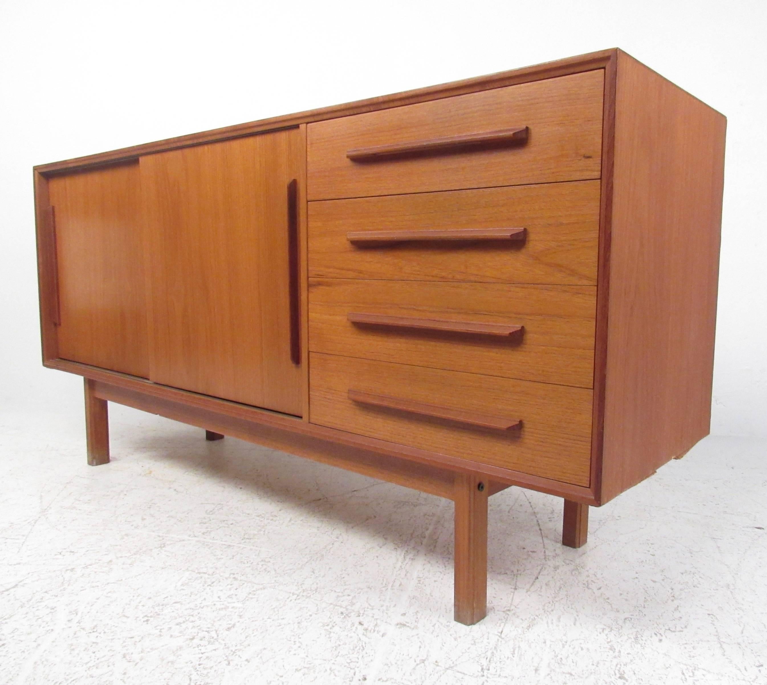 This uniquely sized sliding door credenza features a beautiful Mid-Century teak finish with impressive carved pulls. Four drawers and a spacious shelved cabinet provide plenty of storage in this petite cabinet, ideal for use as a TV console or