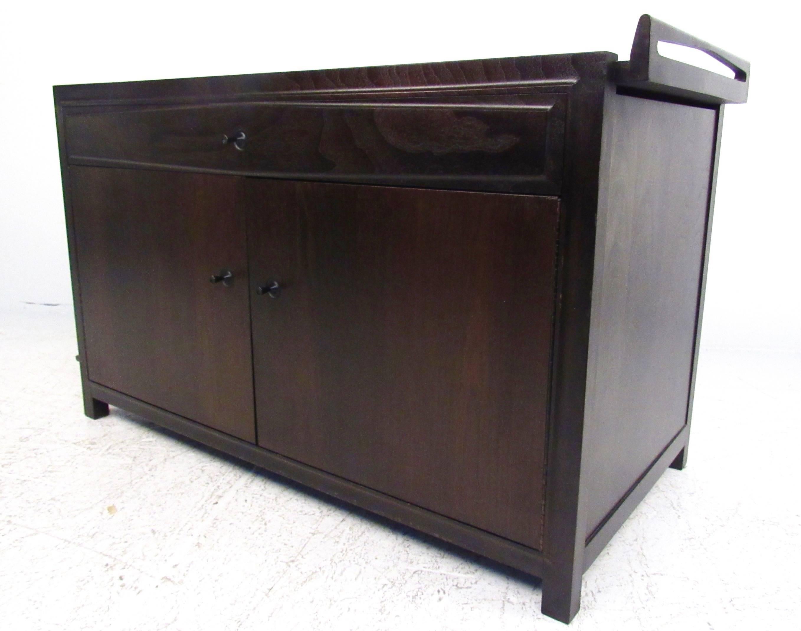 This unique Mid-Century bar cabinet features an expandable formica top with raised edges, spacious storage shelves, and a wide drawer for organizing glass ware or other items. Unique door pulls and dark wood finish add to the vintage appeal of this