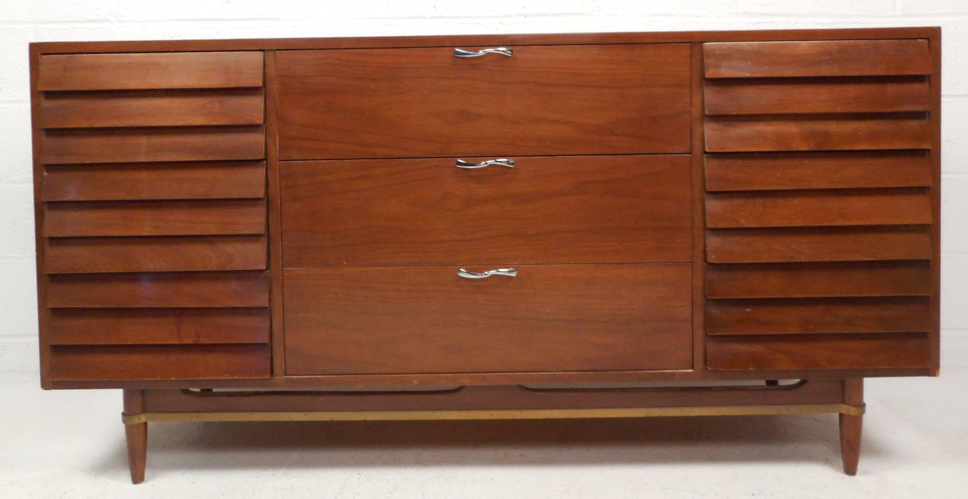 Impressive Mid-Century Modern American walnut dresser features nine hefty drawers providing plenty of room for storage. Sleek design with a sculpted base, brass wraparound trim, and tapered legs. The stylish louvered drawers on the outside and
