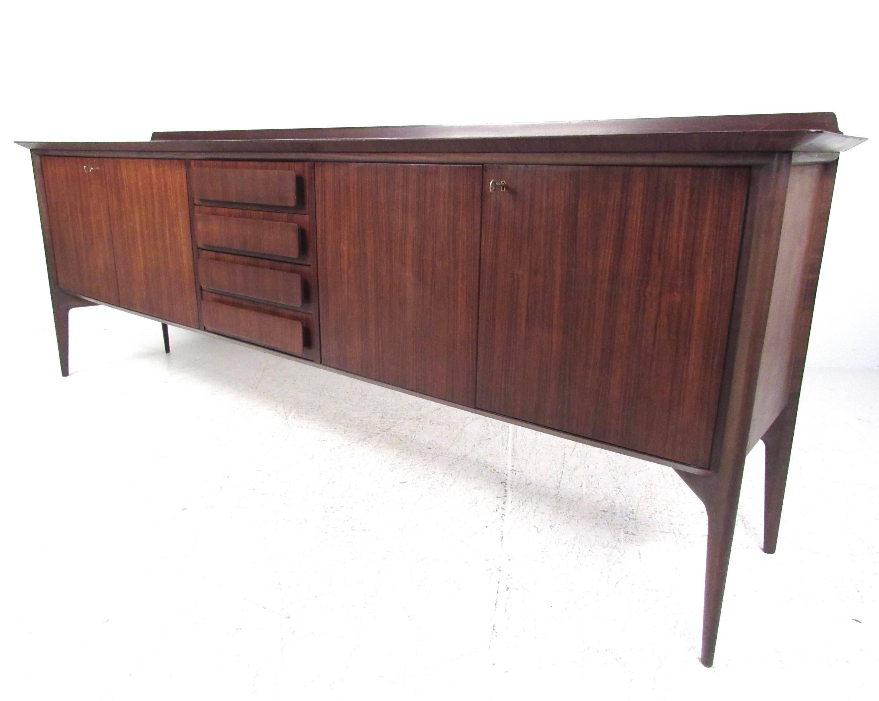 This stunning vintage mahogany sideboard features a dark wood finish with spacious locking cabinets. Raised rear edge, tapered legs, and unique carved front center drawers add to the Mid-Century appeal of the piece. The oversized design of this