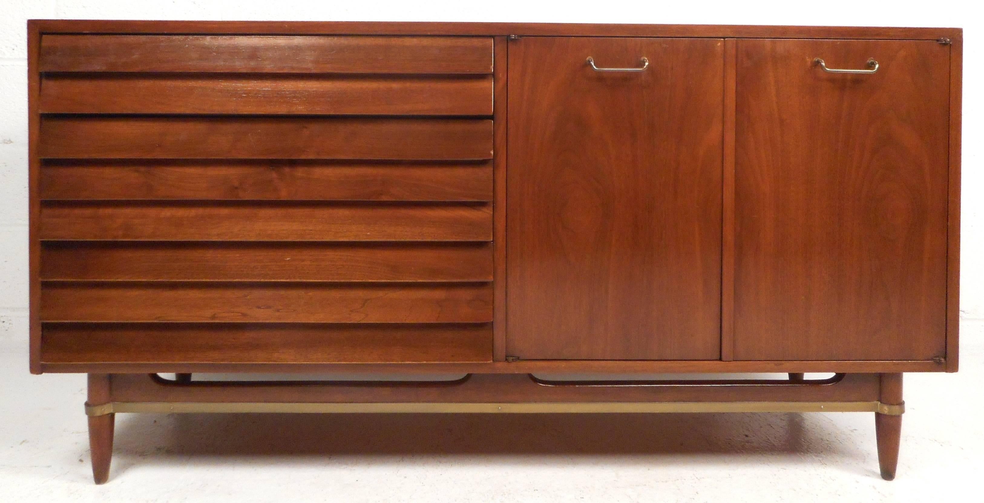 This elegant vintage modern credenza features three large drawers with louvered fronts and three smaller drawers along with a shelf hidden by cabinet doors. Sleek design with unique brass pulls, a brass stretcher, and a sculpted base. Beautiful