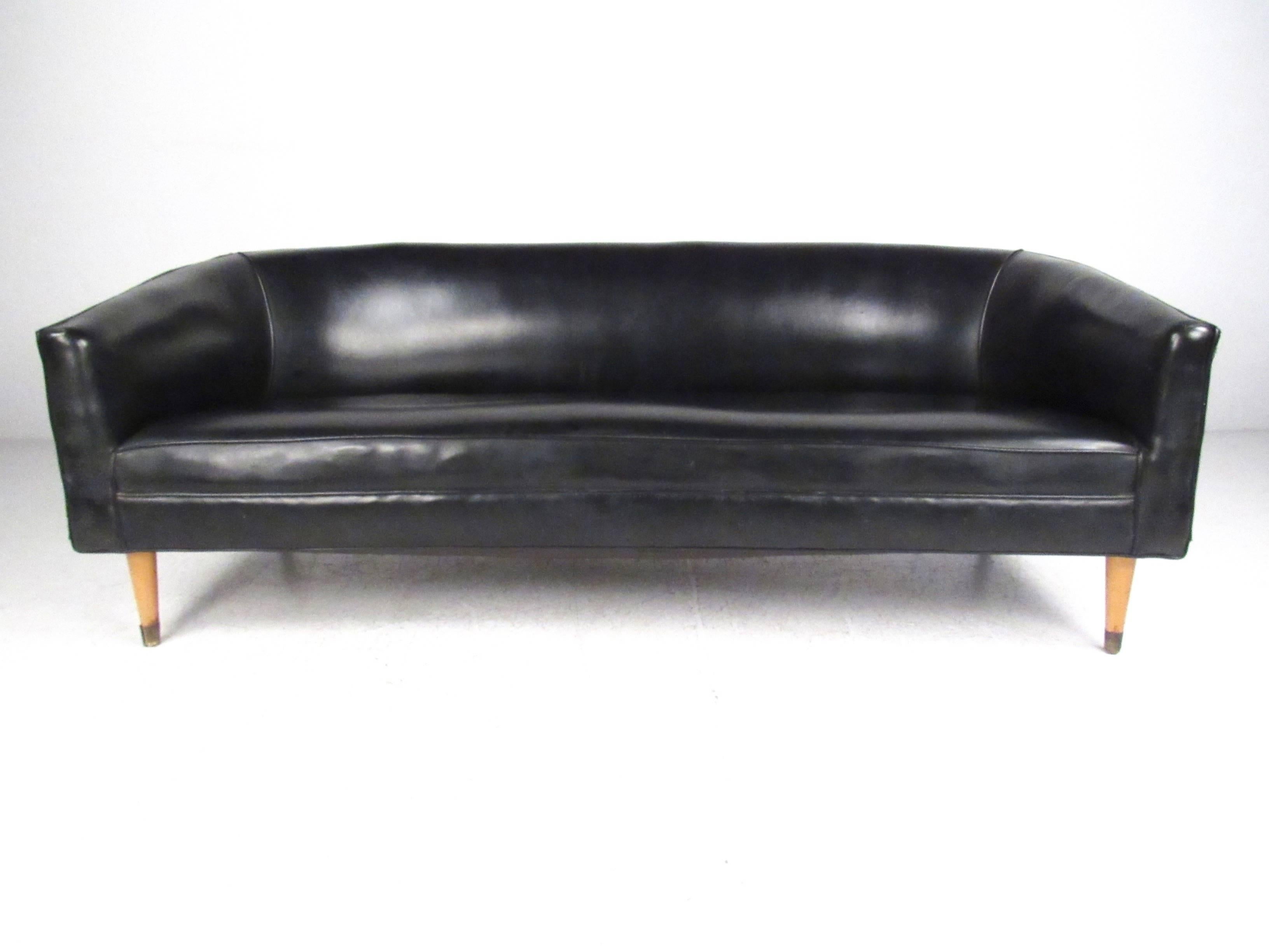 This vintage black Naugahyde sofa features a stylish sounded back design with tapered hardwood legs. Brass trim on the feet and the comfortable design of this Mid-Century piece makes this Italian sofa a stylish addition to any interior. Please