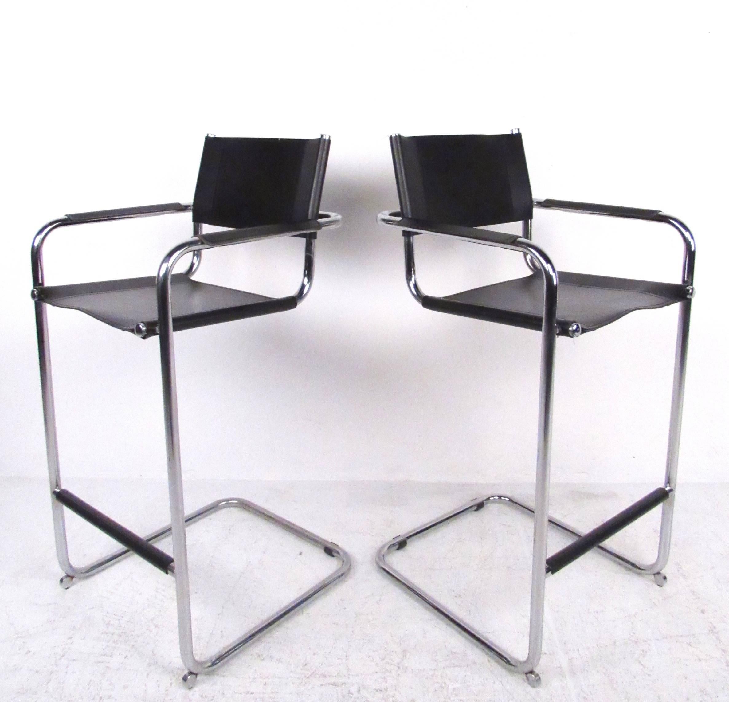 This stylish pair of Stendig style bar stools feature leather strap seats on tubular chrome frames with cantilever bases. Leather arm and footrest adds comfort and Mid-Century style detail to this matching pair. Please confirm item location (NY or