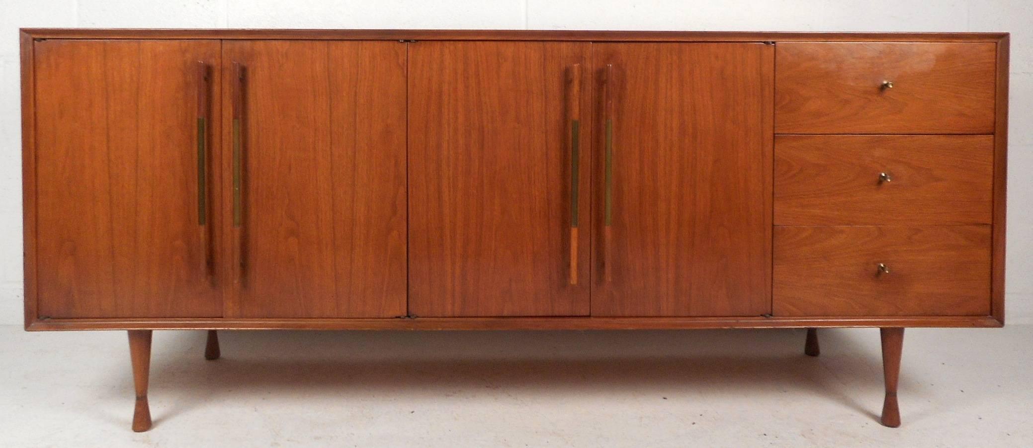 This elegant vintage modern sideboard features unusual long cylindrical pulls on each cabinet with a brass grip in the middle. There is plenty of room for storage within its three large drawers and four cabinets that hide shelving compartments.