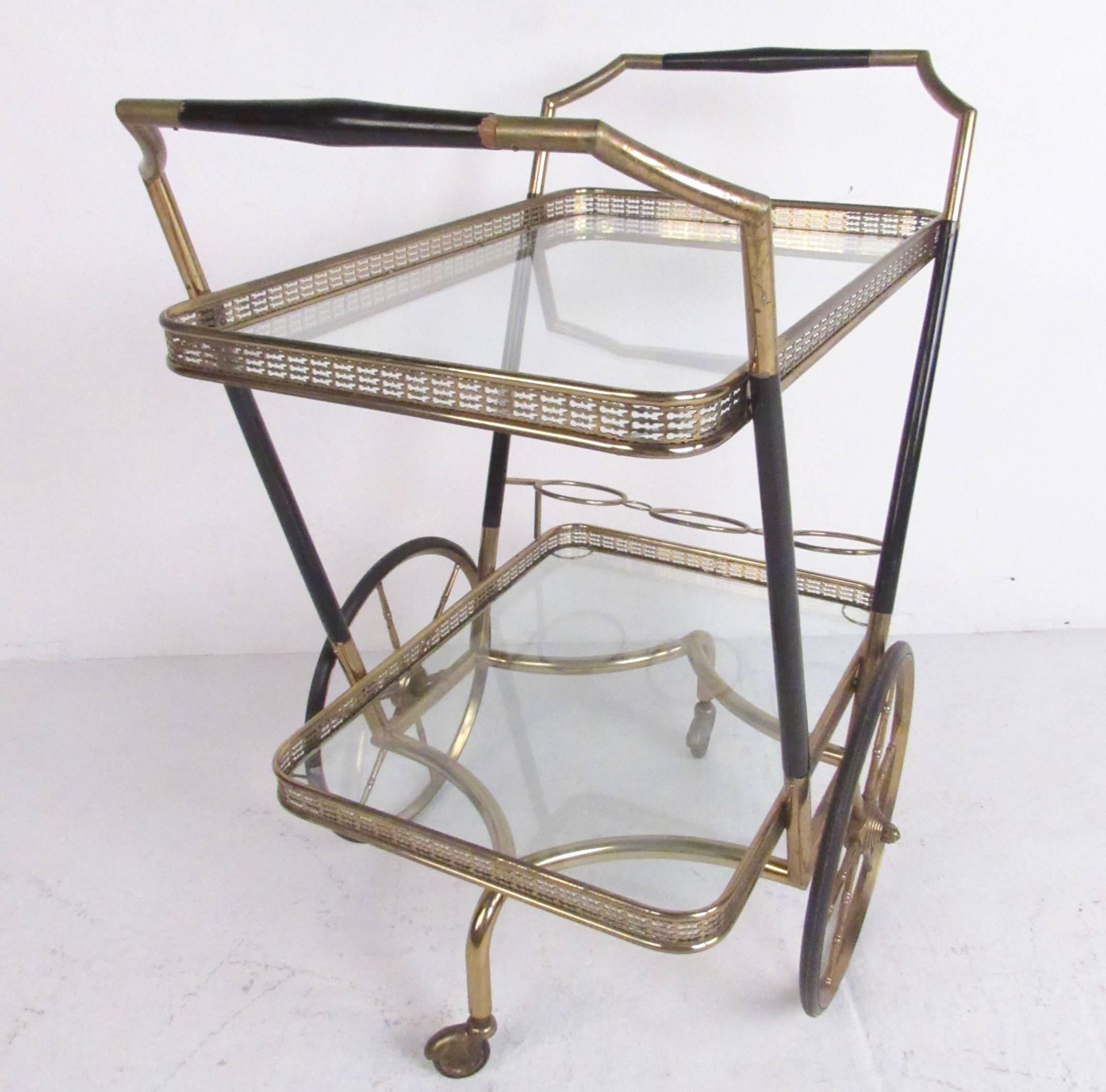 This stunning vintage service cart offers two tiers for storage and display, including handle for use on either side. Ornate trim, brass finish, and ebonized frame add to the Italian modern appeal of the piece, while addition bottle holders make