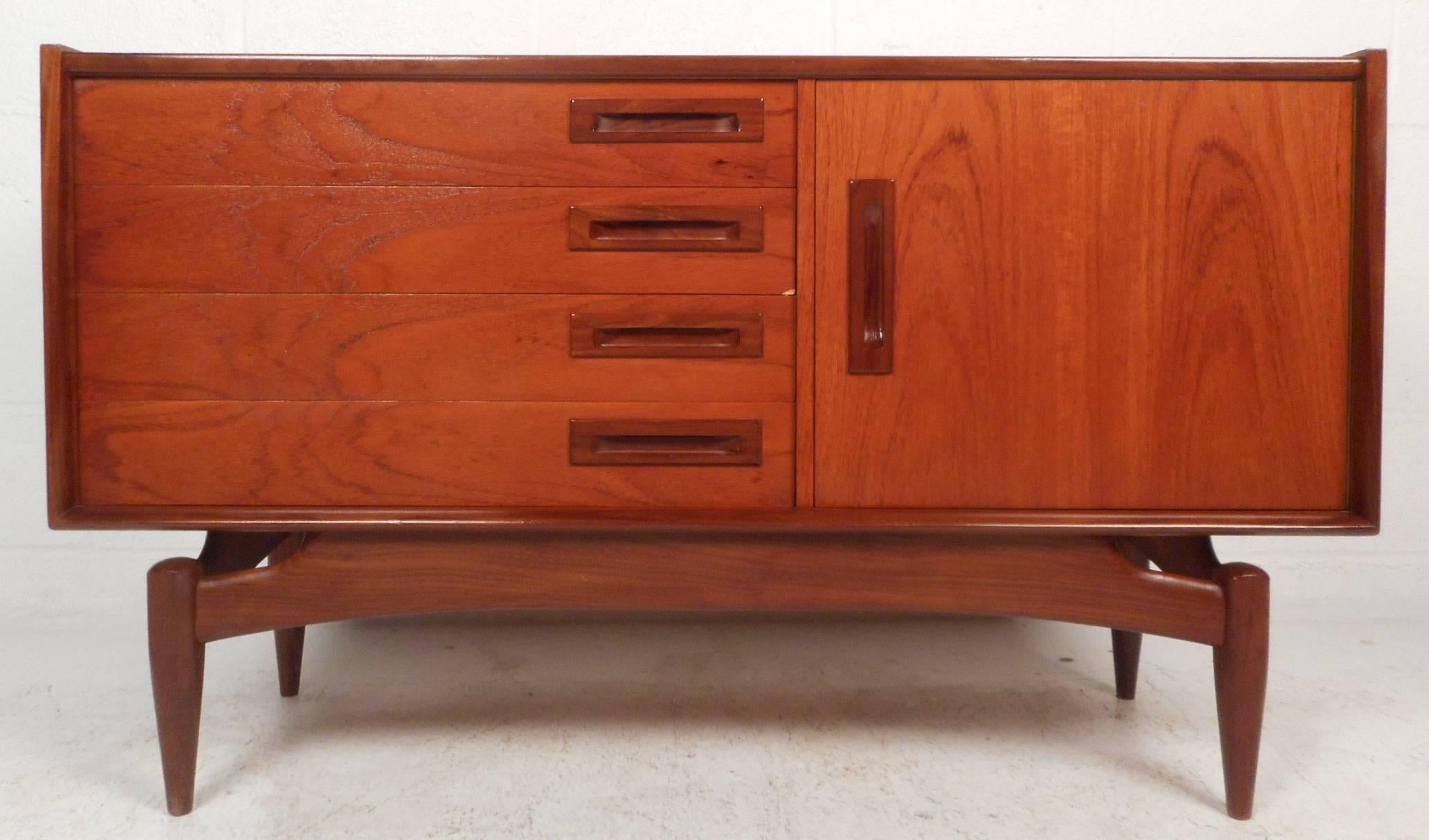 This stunning vintage modern small sideboard features a sculpted base with tapered legs. The stylish design offers plenty of room for storage in its four large drawers and compartment. Unique carved rectangular pulls and elegant wood grain add to