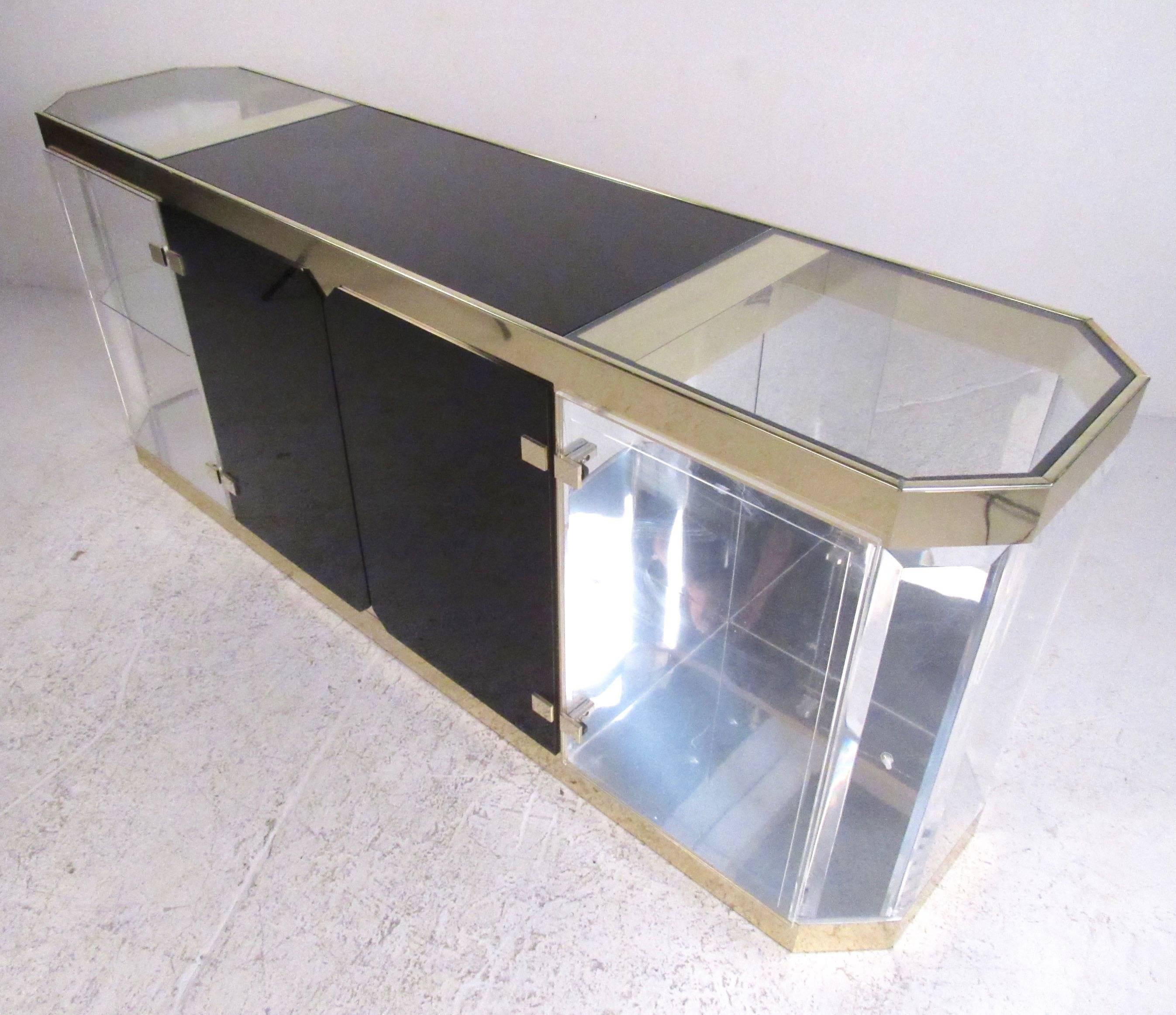 This unique vintage modern display sideboard features stylish brass finish trim, dark glass centre cabinet, and mirrored back Lucite displays on either side. Interior lights add to the value of this sideboard as a showroom display case, as it makes