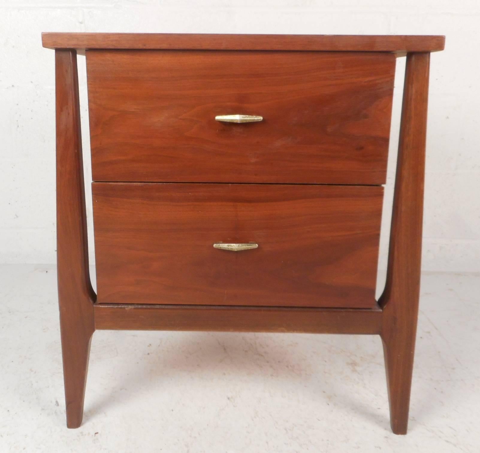 Beautiful vintage modern nightstand features a floating design with angled and tapered legs. Sleek Mid-Century piece has two large drawers with unusual sculpted metal pulls. This end table shows quality craftsmanship with elegant walnut wood grain