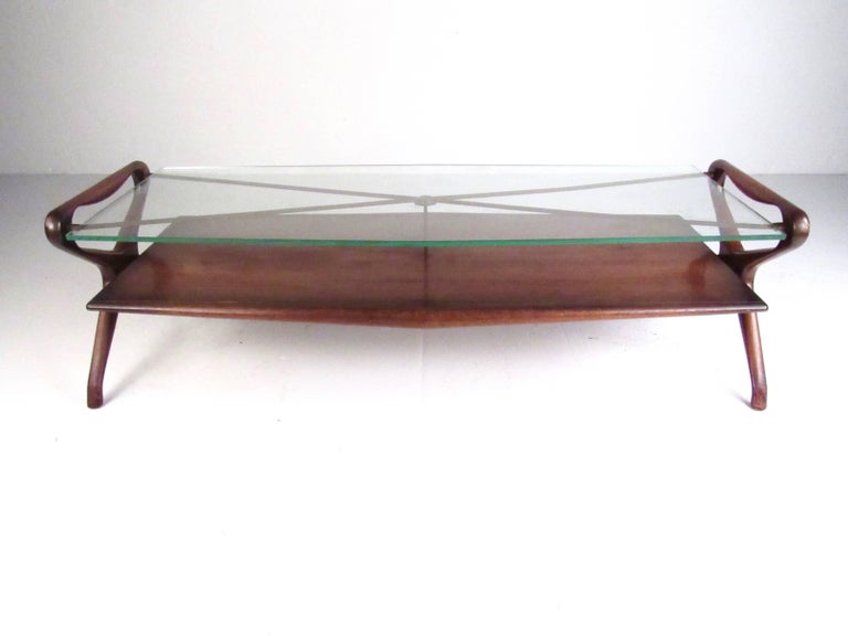 This stunning vintage modern coffee table features unique sculpted wood frame, brass finish details, and thick glass top. Two-tier construction allows for added storage and display, while the Italian modern style of the piece is reminiscent of