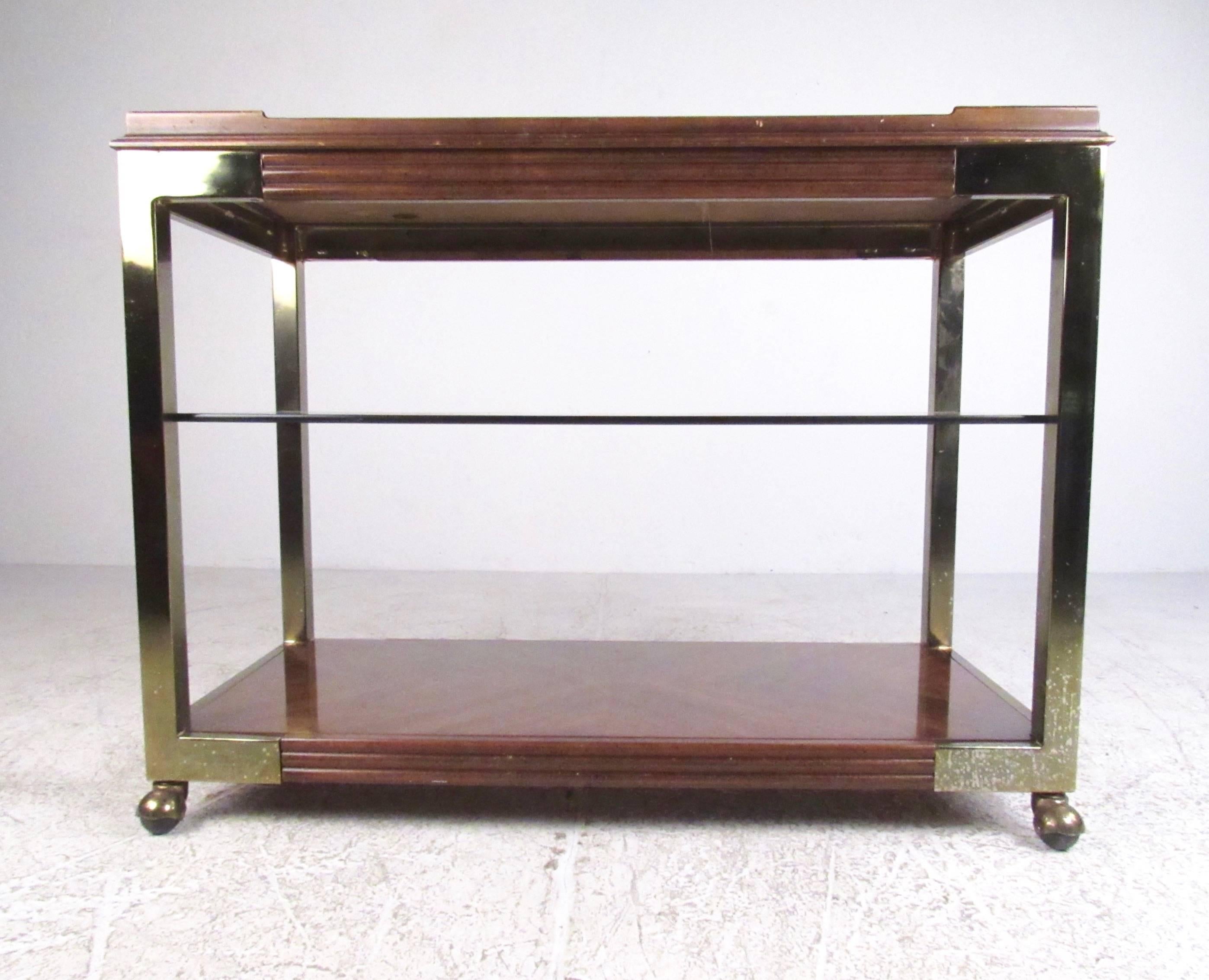 This unique vintage modern service cart features two-tier design allowing for plenty of liquor and glassware storage for home or office. Dark wood finish with brass frame nicely compliments the bronze mirrored top finish and tinted glass shelf,