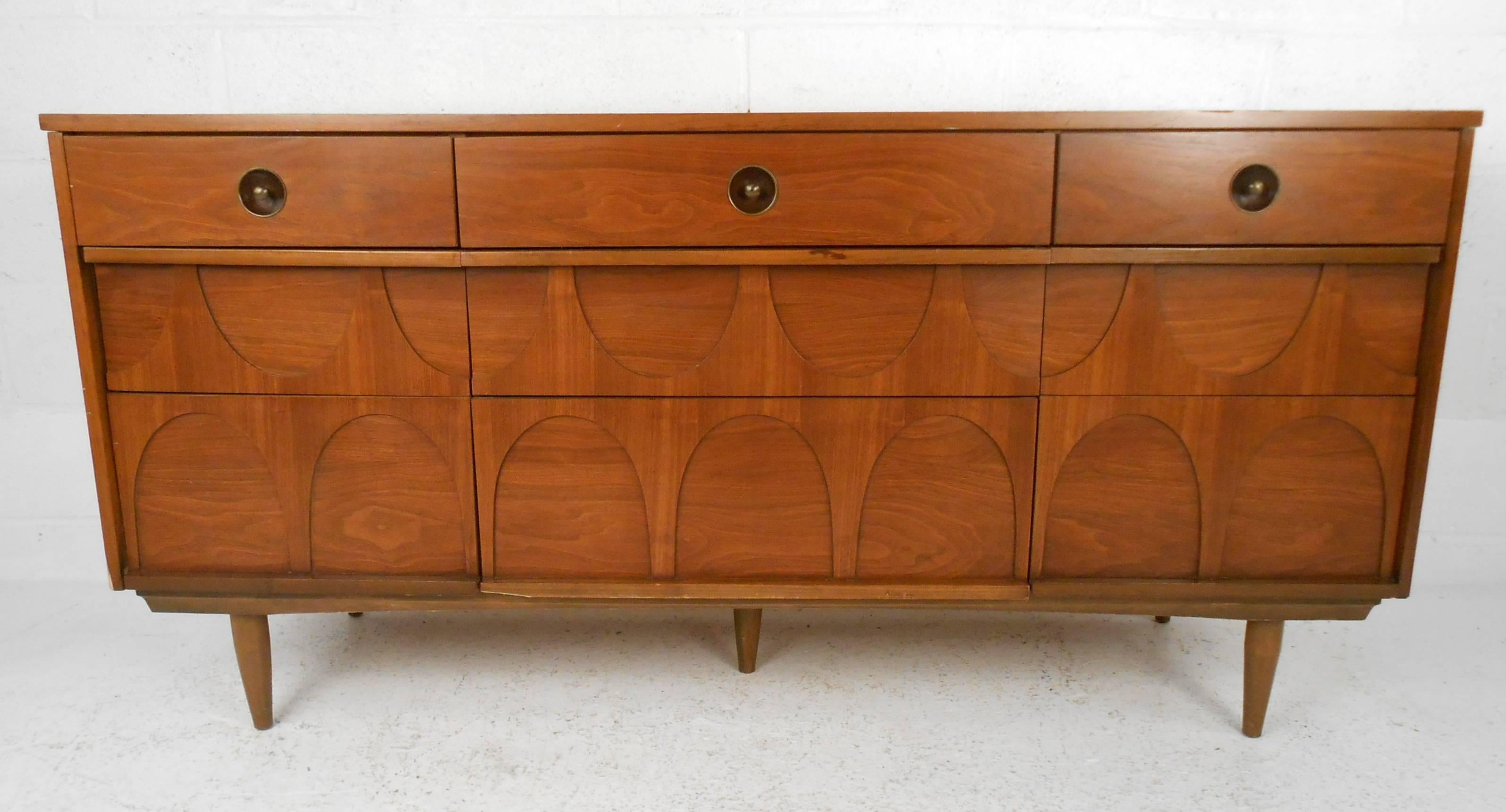 This beautiful vintage modern dresser features nine large drawers and a sculpted front. Unique circular recessed drawer pulls, elegant walnut wood grain, and tapered legs add to the allure. Quality craftsmanship with a centre leg for added