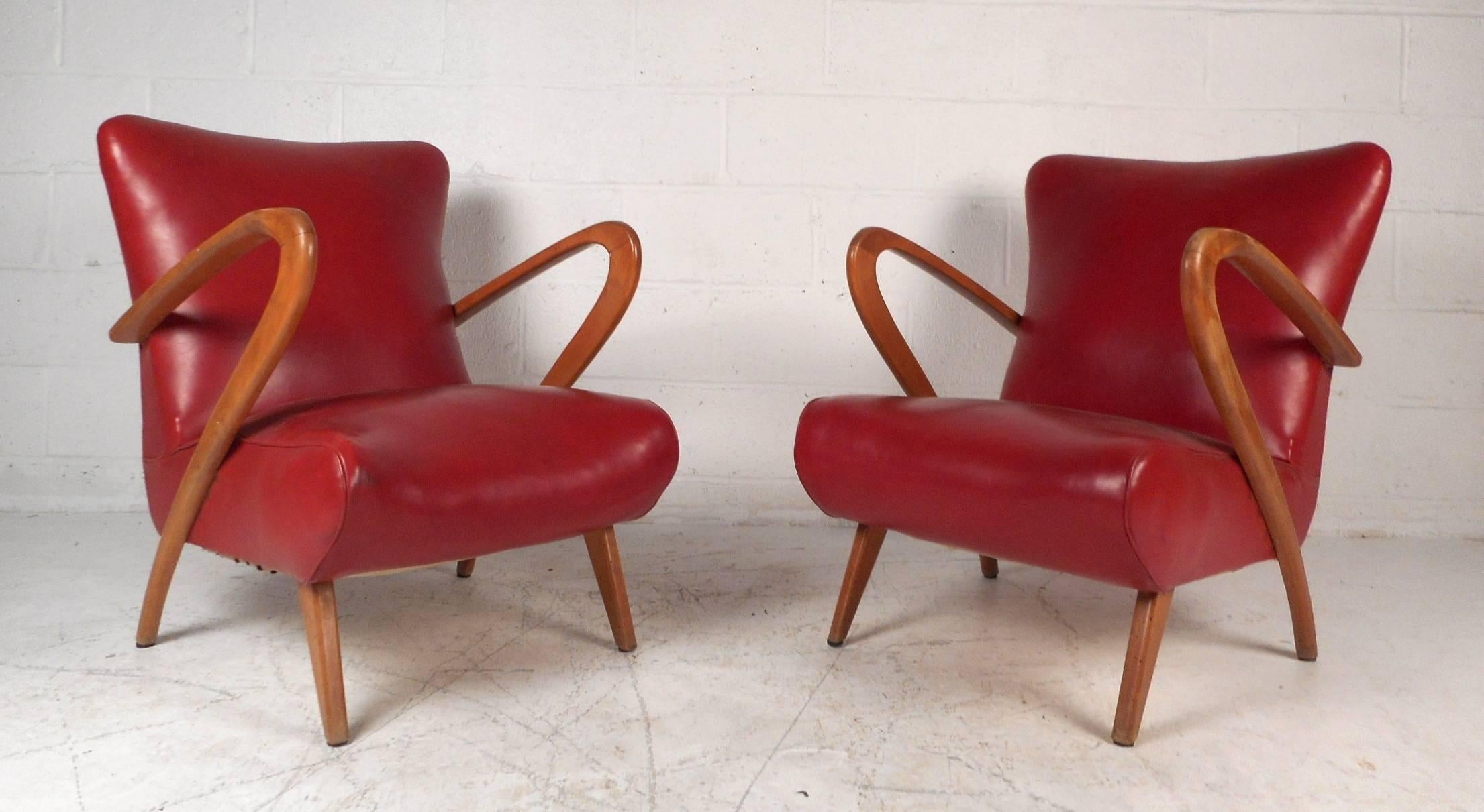 This beautiful vintage modern pair of lounge chairs feature sculptural armrests, angled legs, and elegant light wood grain. This comfortable pair have thick padded seating and elegant red vinyl upholstery. Sleek design has the perfect contours for