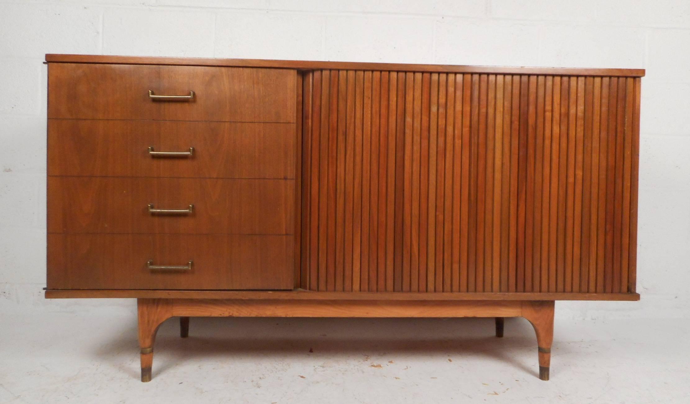 This gorgeous vintage modern credenza features six hidden drawers behind a cabinet and a tambour door. Unique cabinet door has appearance of four faux drawers with sculpted metal pulls. Sleek compact design has tapered legs with brass trim around