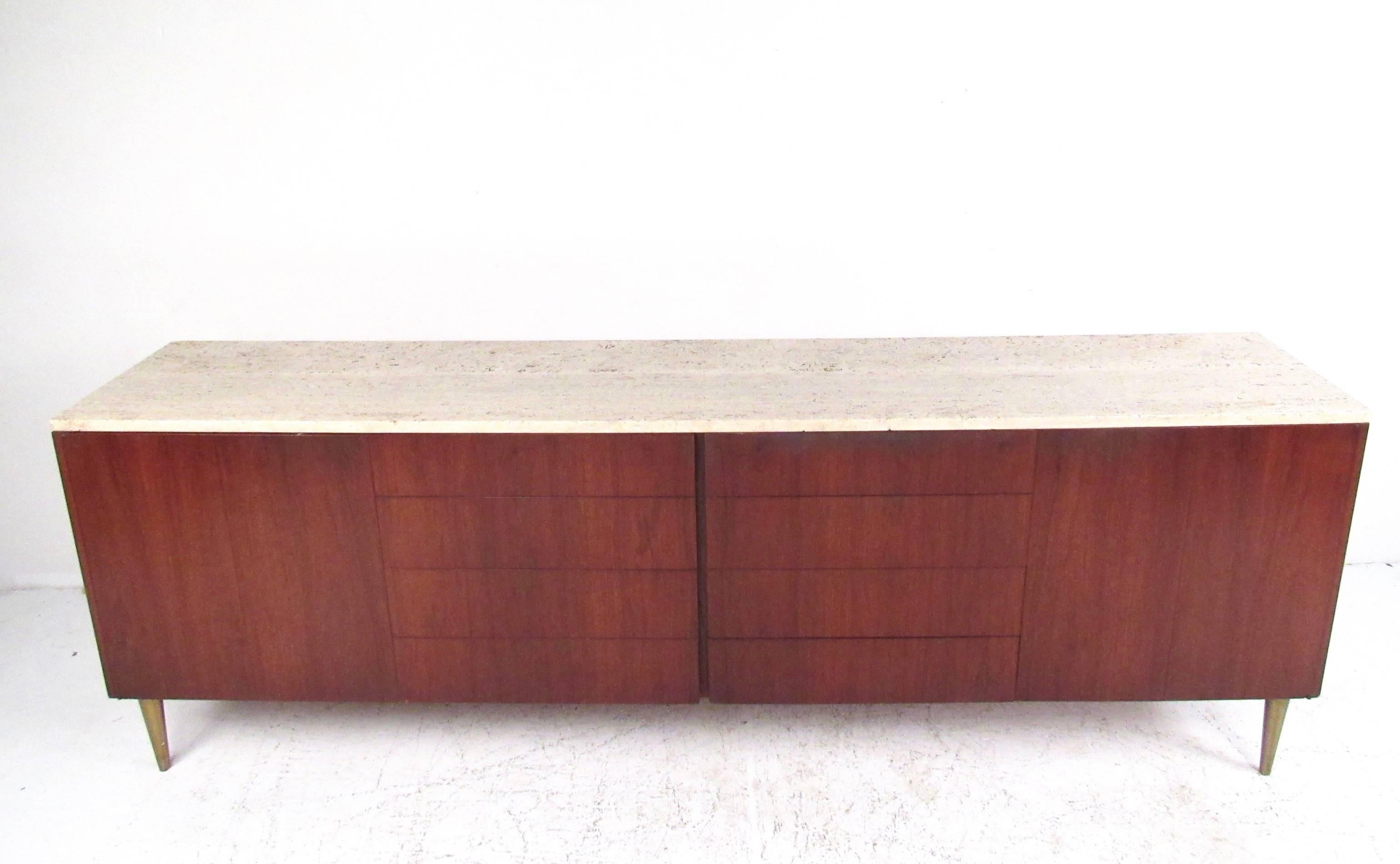 Stylish long midcentury sideboard features bi-fold doors hide mixed shelf and drawer storage. Unique travertine top compliments the rich teak finish of the piece, while tapered brass legs round out the vintage appeal. Please confirm item location