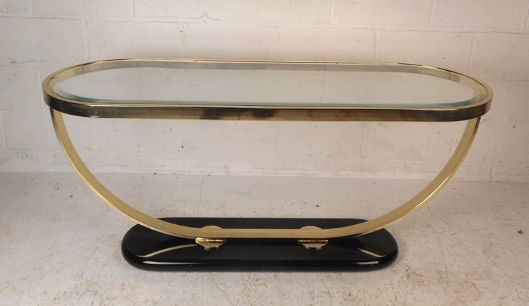 Stunning vintage modern ellipse shaped console table with a black oval base. Sleek design with a brass frame and beveled glass with a light a tint of green. This stylish vintage modern piece looks great behind the sofa, in the entryway, or in the