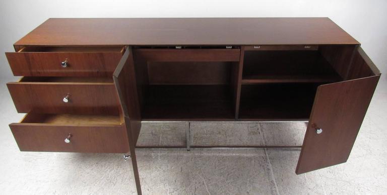 Elegant Mid-Century style server with bi fold doors revealing adjustable shelf storage and felt lined cutlery drawer on right and three storage drawers on left.
Chrome square tubular base and matching chrome pulls compliment the dark tone wood