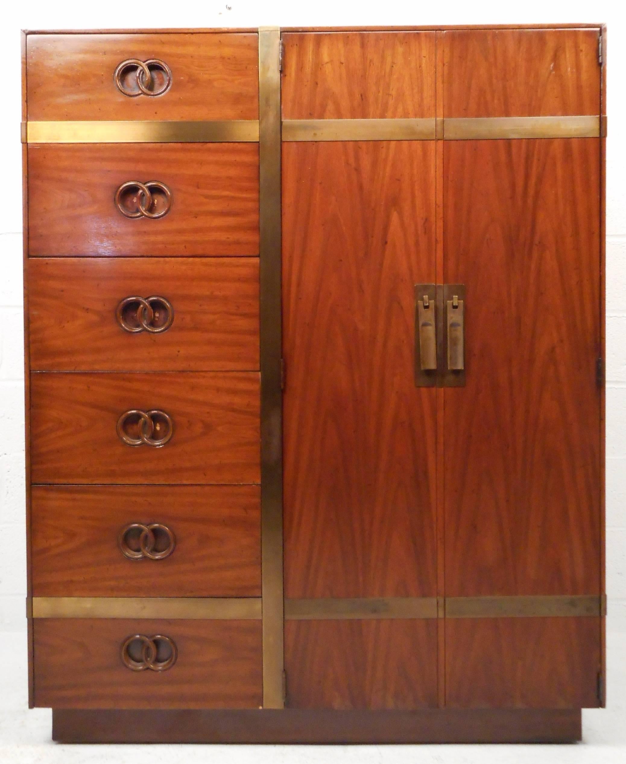 Impressive Mid-Century Modern wardrobe by Heritage features vintage walnut finish, unique interlocking circular pulls, brass trim and brass cabinet pulls. The elegant design provides a mix of cabinet and drawer storage space, while making a stylish