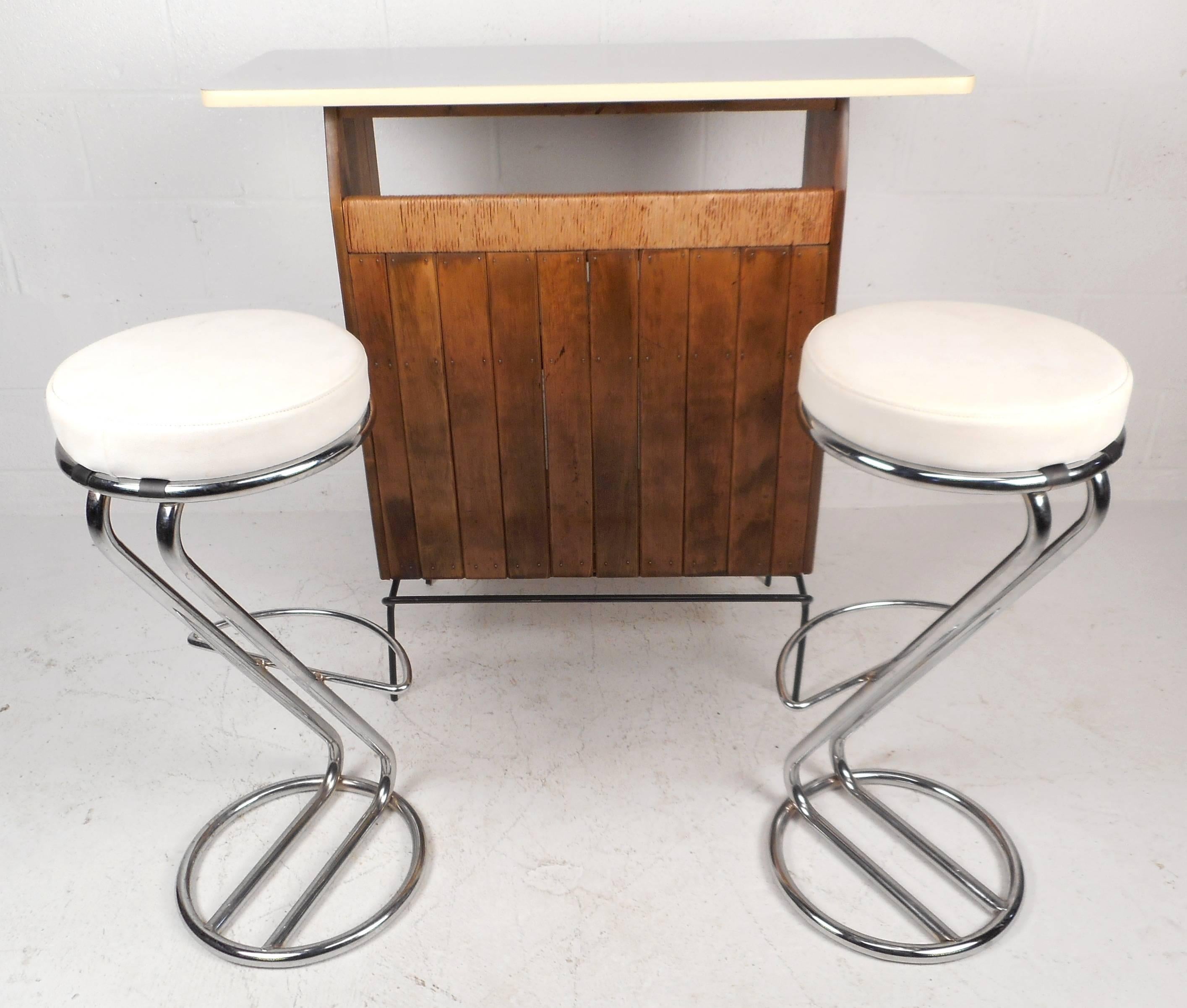 This vintage modern compact dry bar features beautiful raffia detail and open wrought iron framework with a footrest. The sleek bar has a slatted oak front, white formica top and two rear shelves for storage. The versatile design can be used as a