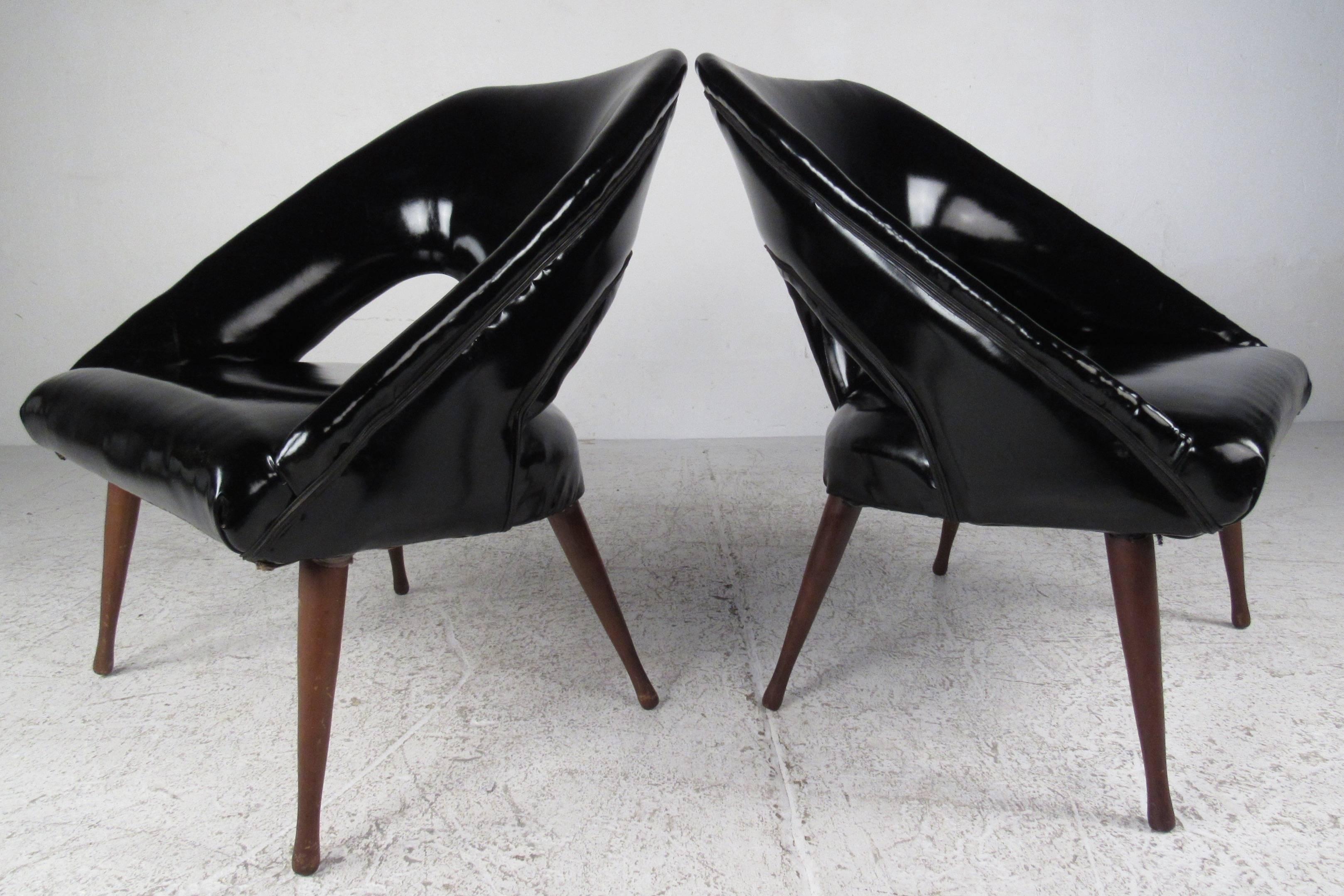 Sleek pair of vintage modern glossy black vinyl lounge chairs with tapered wood legs. This unusual pair of chairs feature a barrel back rest with two cut outs and low angled arm rests. Thick padded seating and splayed drumstick legs make this