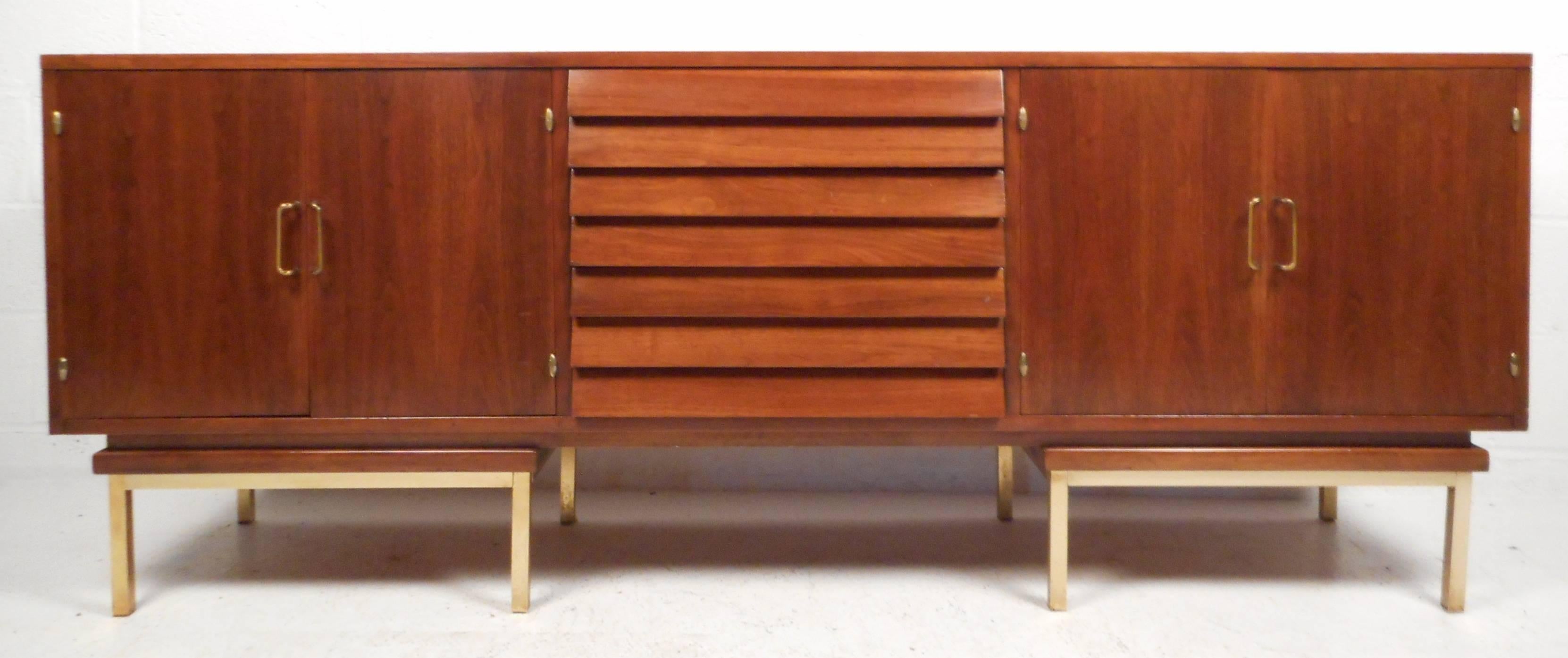 This massive vintage modern sideboard features three louvered drawers in the middle and two large storage compartments on the sides. The rich walnut finish, sculpted brass handles, and unique brass base make this piece an impressive addition to any