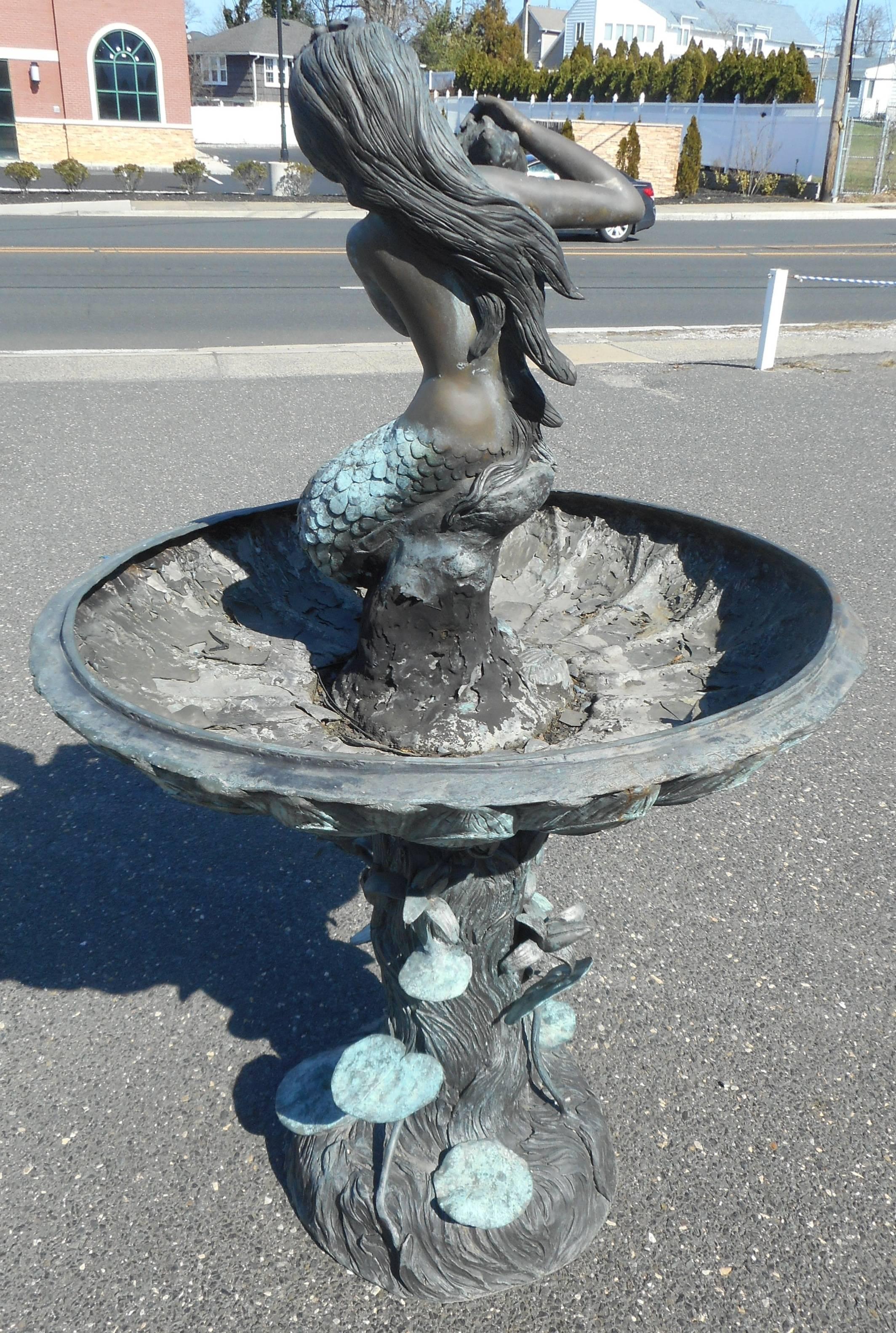 This beautiful bronze mermaid fountain features a mermaid perched on the top with a shell that spills water into the reservoir below. The unique base resembles a mound with flowers and lily pads. Elegant design with intricate detail on beautifully