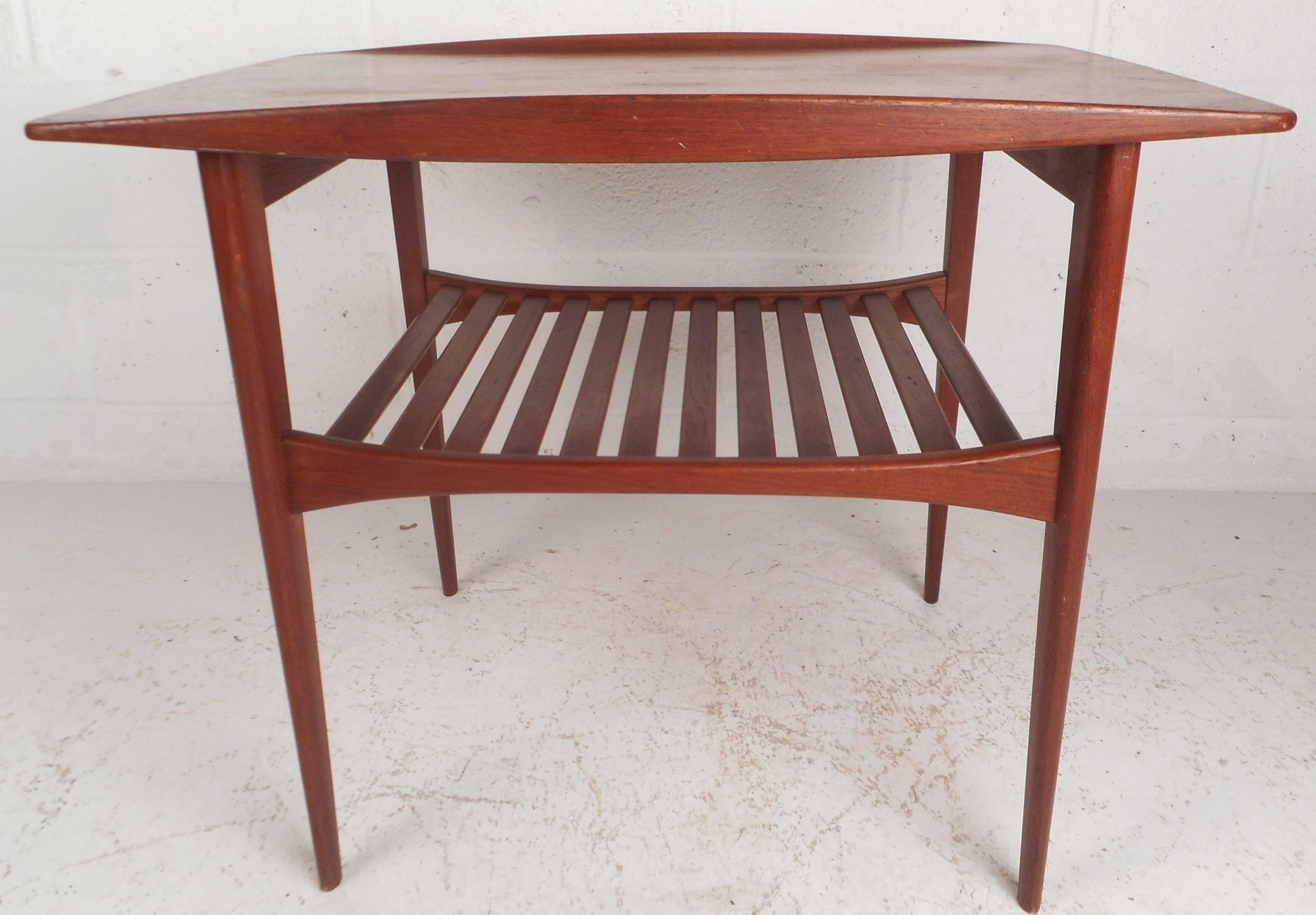 This beautiful vintage modern end table features two tiers for storage and stylish tapered legs. The sleek design has unique raised edges on the top and a slatted lower tier. Quality construction with elegant teak wood grain makes this Mid-Century