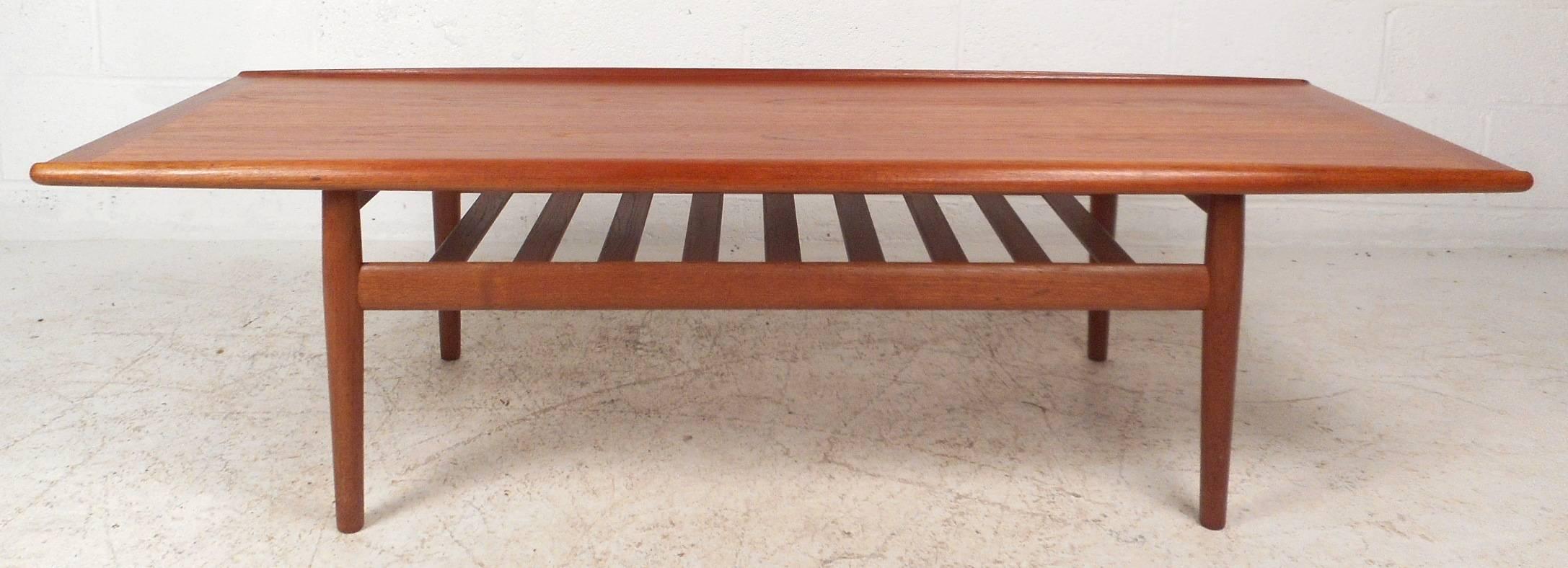 This beautiful vintage modern coffee table features unique raised edges on top and stylish tapered legs. The sleek Grete Jalk design has a bottom slatted tier for additional storage. Quality construction with a rich teak finish makes the perfect