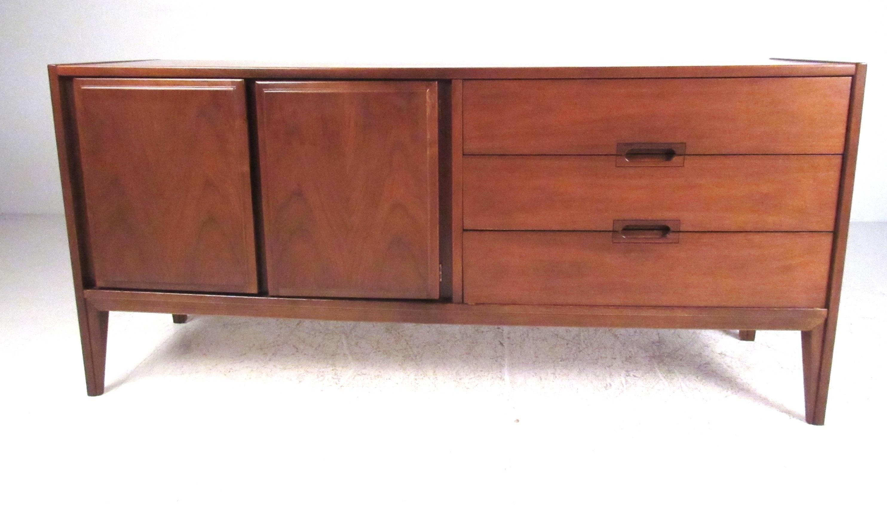 Mid-Century style walnut credenza by United Furniture featuring three drawers with recessed pulls and double door storage with adjustable shelf. Please confirm item location (NY or NJ) with dealer.