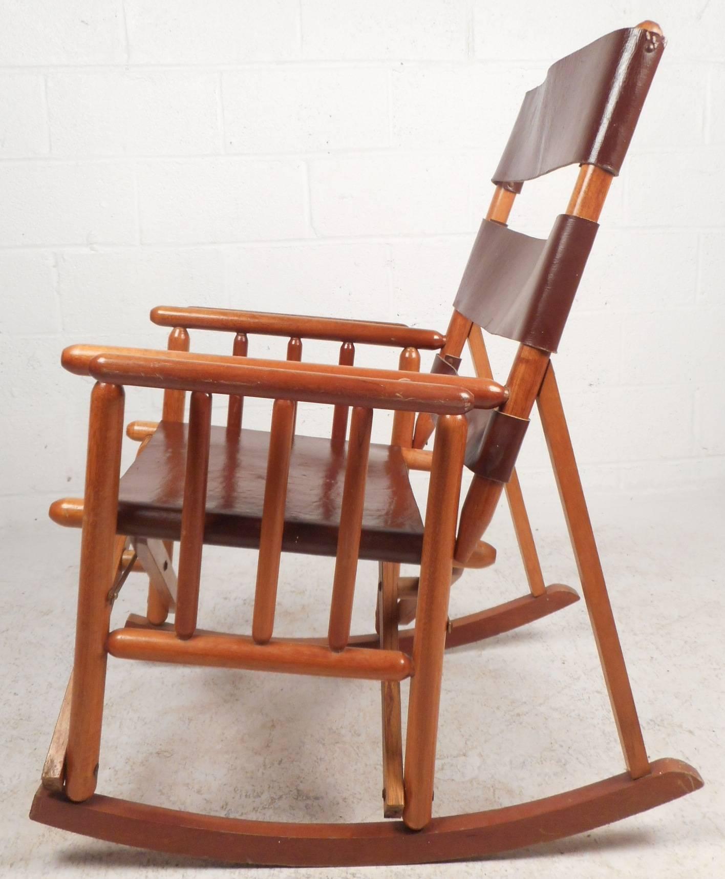 Beautiful vintage modern rocking chair with three thick strips of leather on the backrest and a wide leather seat. Sleek walnut cylindrical frame with unique spindle sides and double wide armrests. Versatile lounge chair conveniently folds for easy