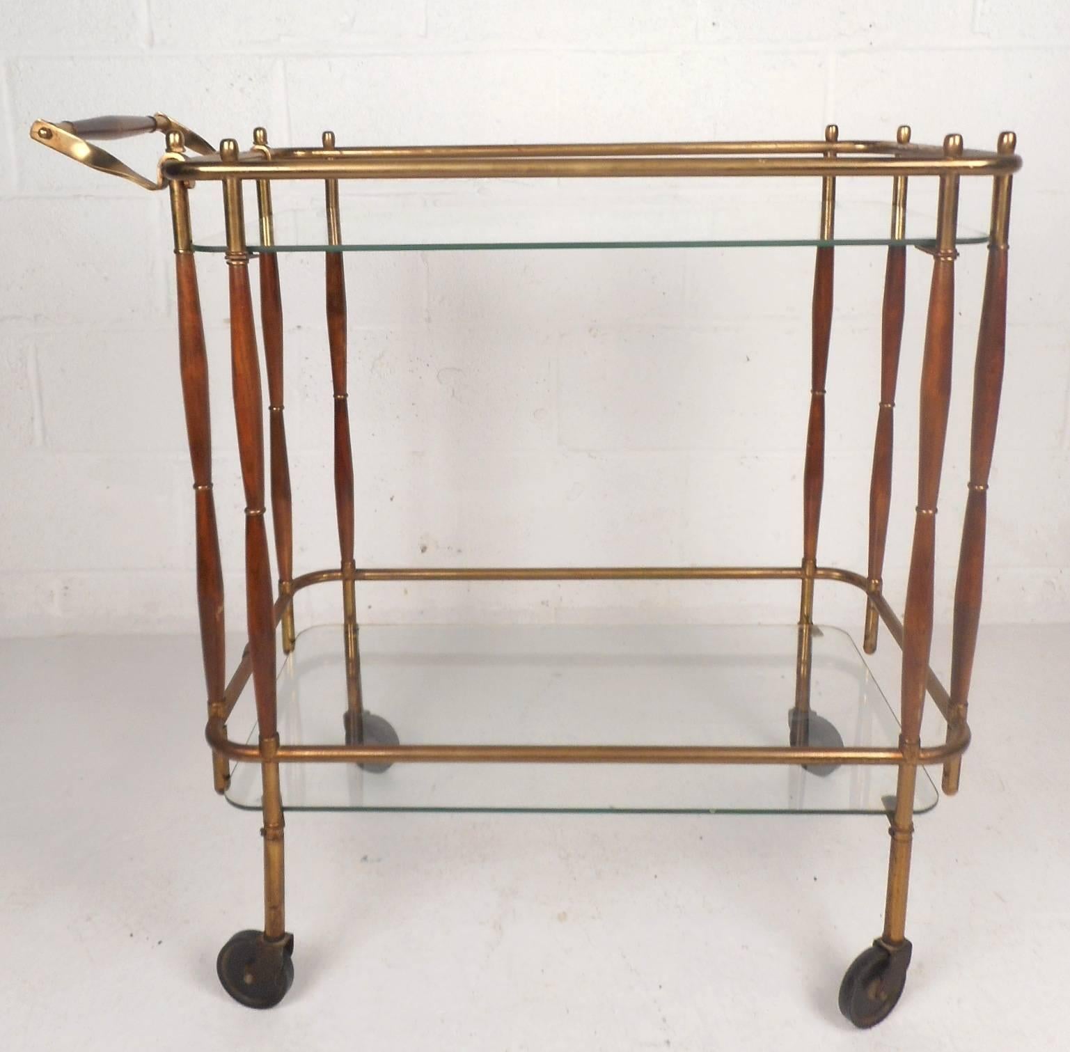 This beautiful vintage modern bar cart features a stylish two-tone brass and wood frame on wheels. Sleek and functional design with two tiers of glass shelving for placing items. Unique design shows quality construction and makes the perfect eye