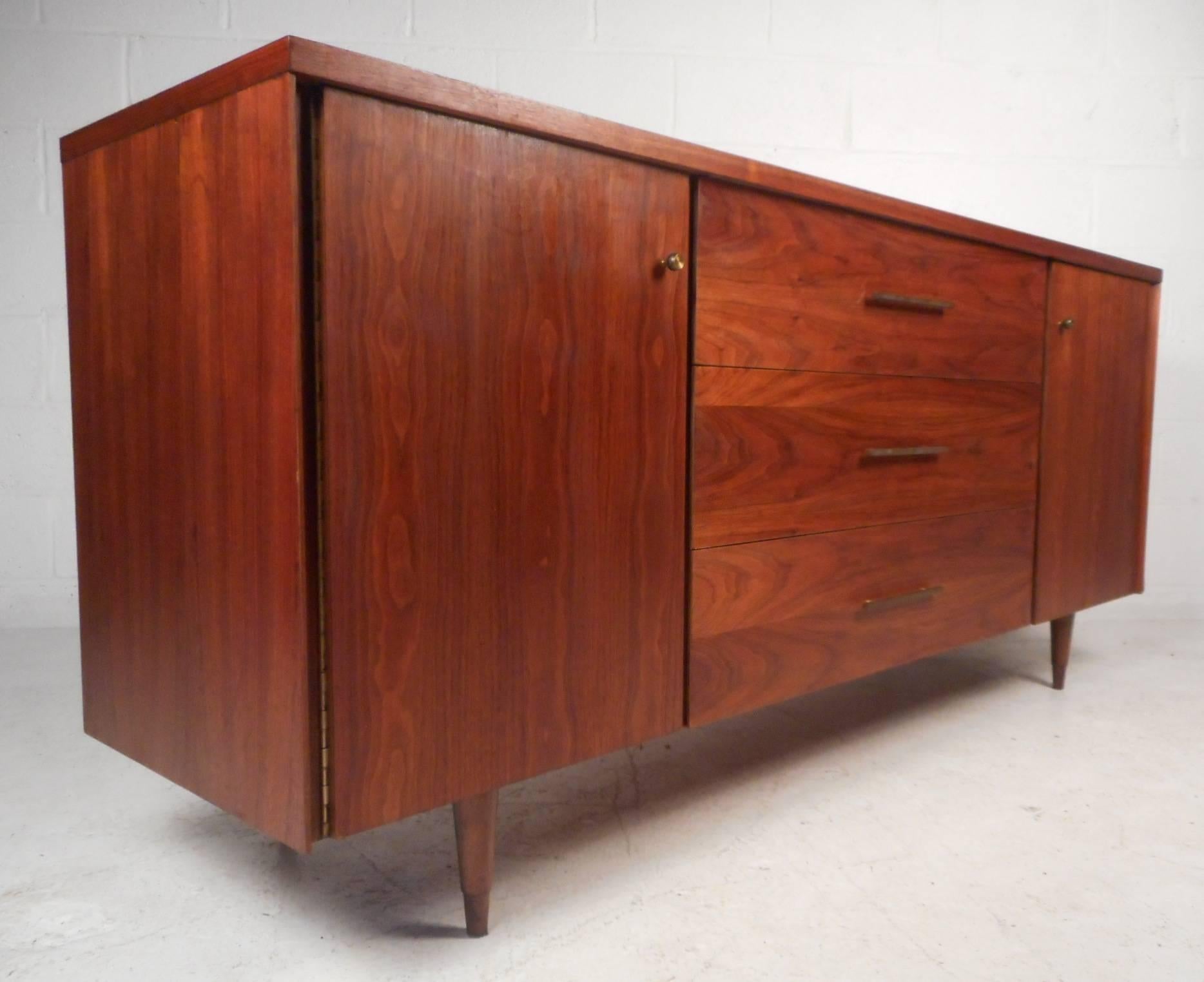 This beautiful vintage modern sideboard features three large drawers and two compartments with shelves hidden behind cabinet doors. Unique brass pulls and tapered legs add to the allure. Quality craftsmanship with elegant walnut wood grain