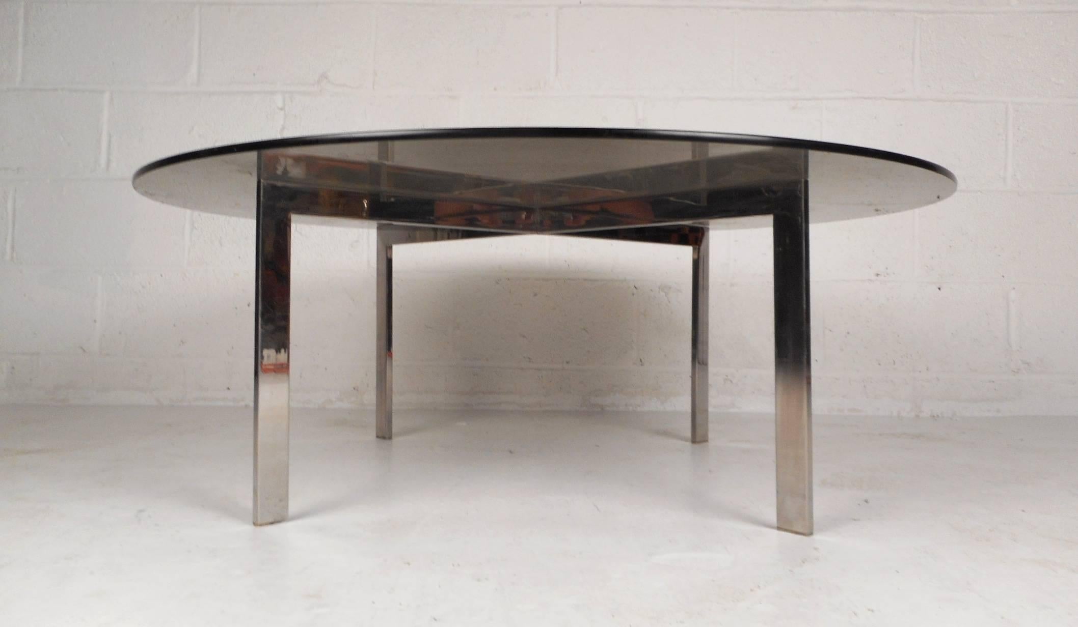 This beautiful vintage modern coffee table features a unique flat bar chrome base with a round glass top. The sleek design has a thick glass top with a smokey tint sitting on top of a heavy "X" shaped chrome base. This wonderful midcentury