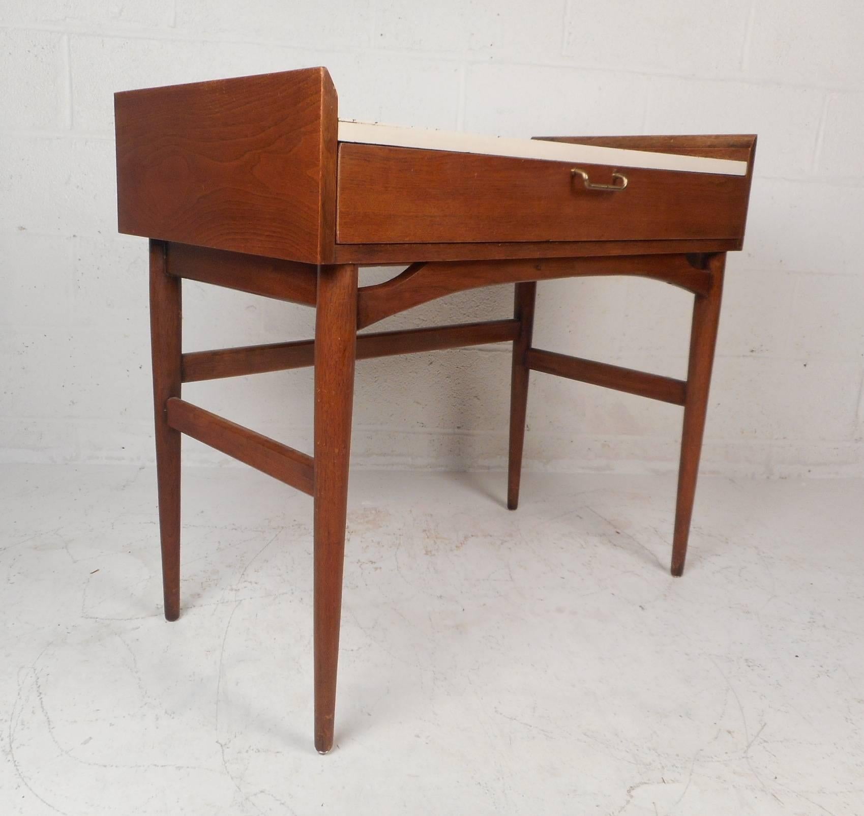 This beautiful vintage modern vanity features one drawer with a unique brass pull and slender tapered legs. Sleek design with raised and beveled wood edges surrounding the stylish white formica top. Quality construction with stretchers running to
