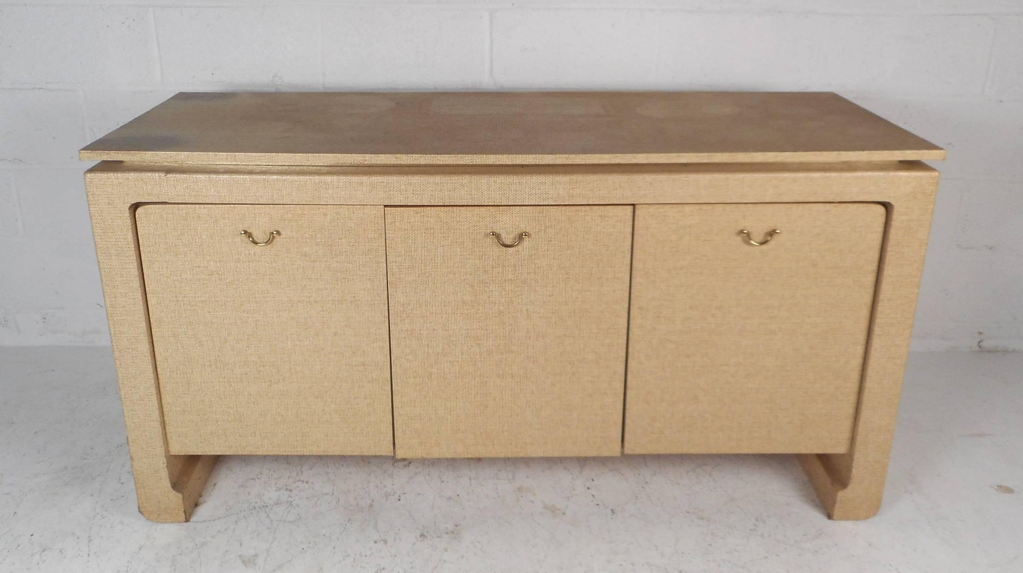 This beautiful vintage modern sideboard features three cabinet doors with unique brass pulls that open up to unveil large compartments with shelves. Unusual design covered in grass cloth with stylish brass trim wrapping around the underside of the