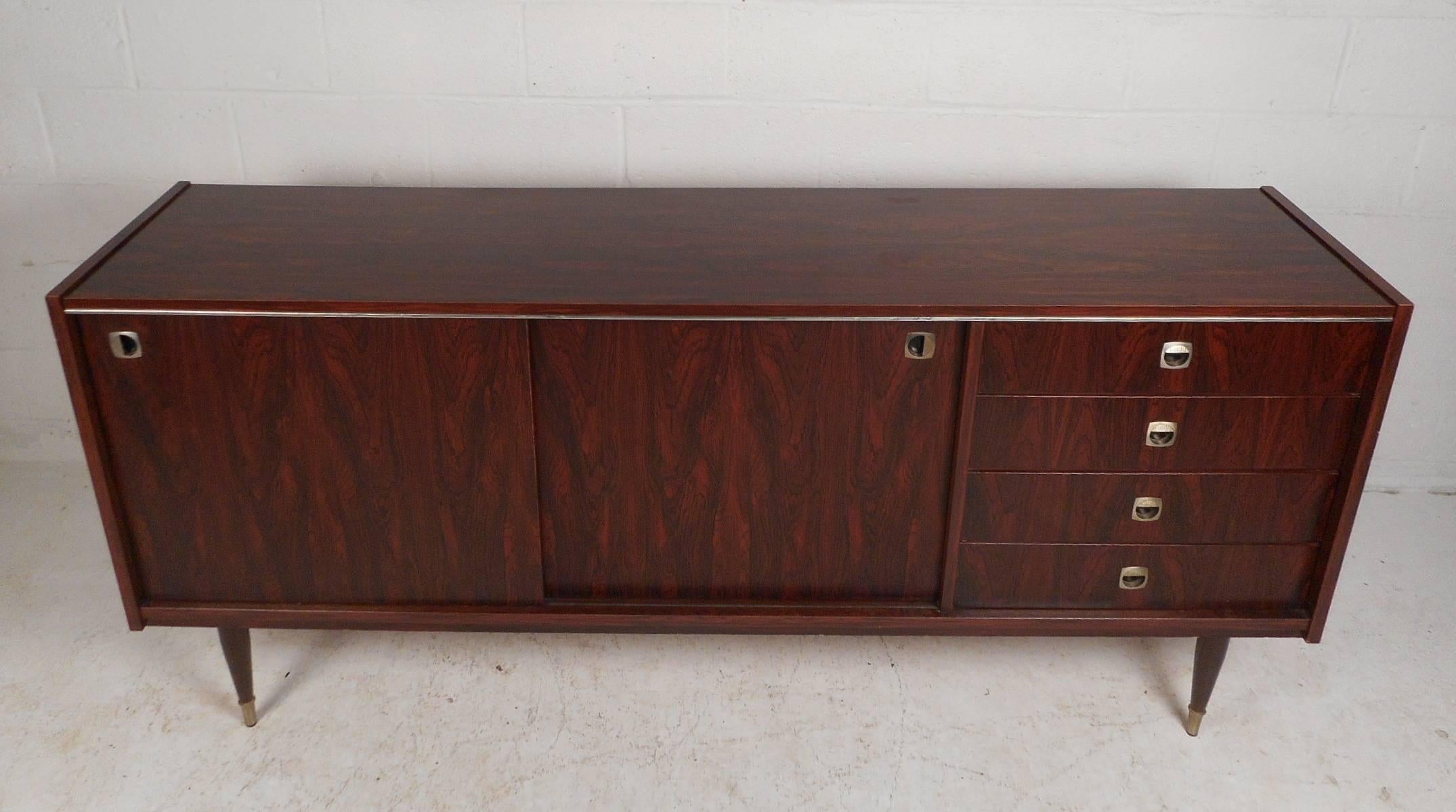 This stunning mid-century modern sideboard offers plenty of room for storage within its four hefty drawers and large storage compartments with shelves hidden behind its sliding doors. An elegant rosewood wood grain throughout and tapered legs with