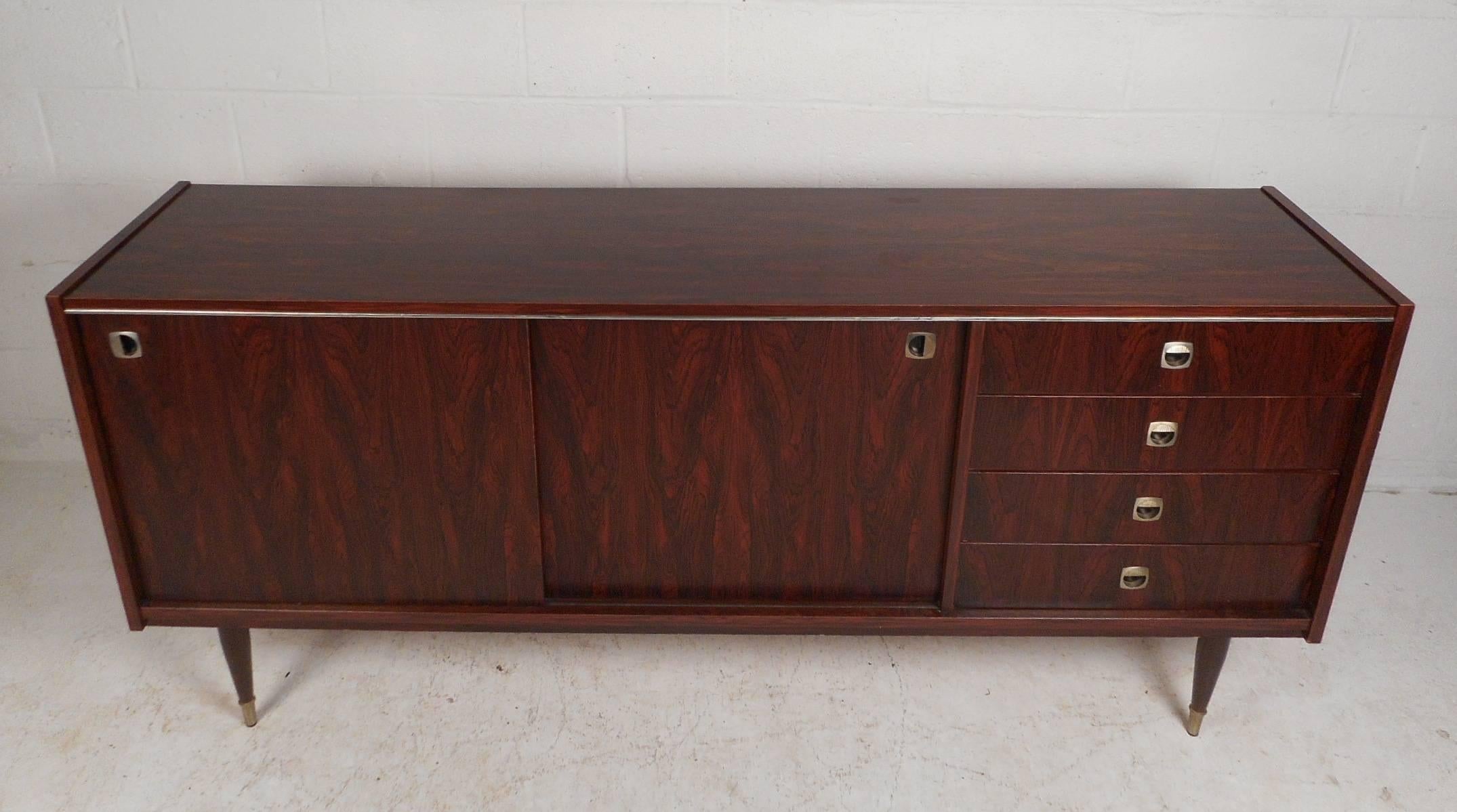 Questa splendida credenza moderna di metà secolo offre molto spazio per riporre i tuoi oggetti grazie ai quattro cassetti e agli ampi vani portaoggetti con ripiani nascosti dietro le ante scorrevoli. L'elegante venatura del legno di palissandro e le