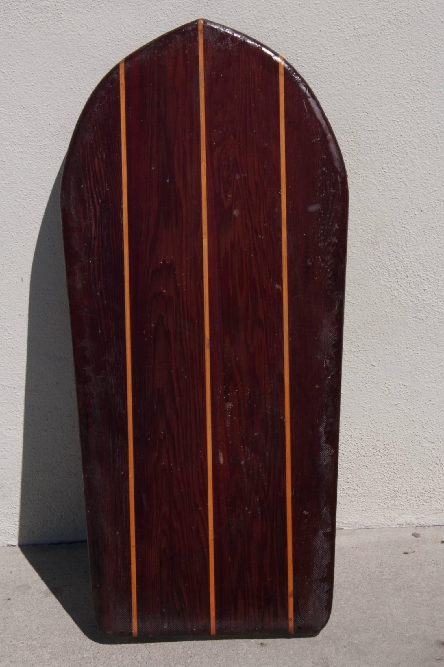 Redwood Twin-fin belly board with Hardwood Stingers, circa 1950.
Dark redwood accented by stripes of light wood stingers captures the eye. Classic shape featuring unique concave bottom and twin fins captures the imagination. Authentic wear
