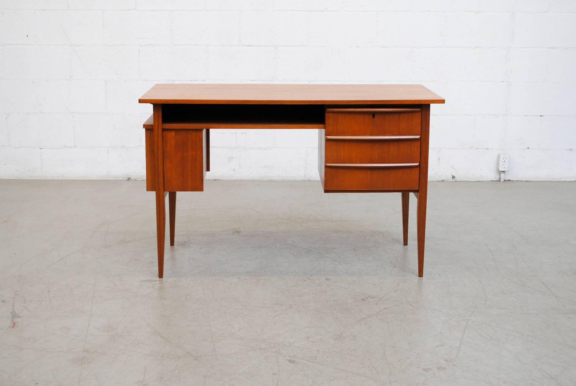 Beautiful Mid-Century teak desk. Three drawers on the right hand side with key lock and side bookshelf. Center cubby hole for storage and tapered legs. Good Original Condiiton.