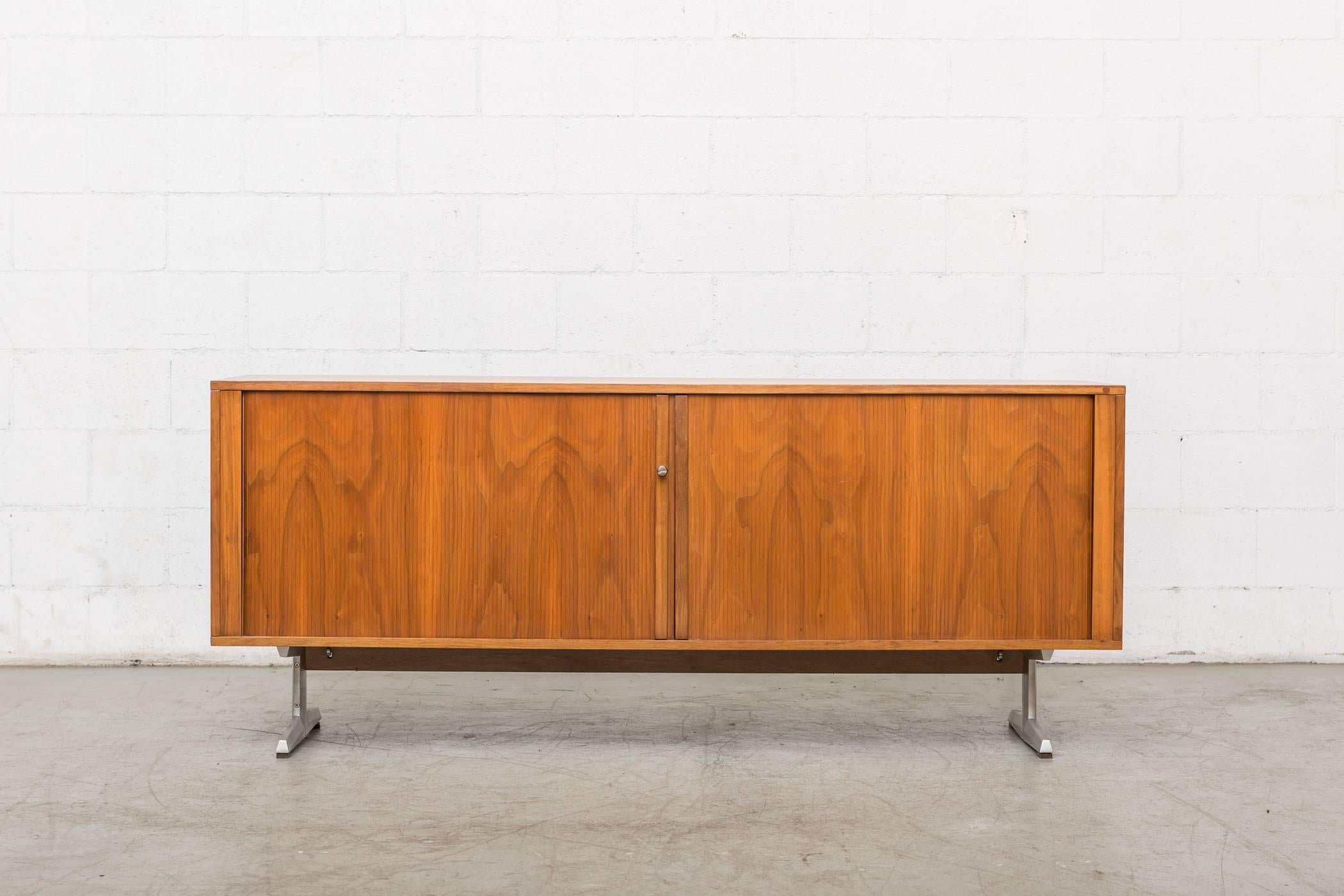 Midcentury teak credenza. Beautiful sliding tambour doors with three sections of interior storage with adjustable shelves. Enameled metal base with chrome T-shaped legs. Good original condition. Key lock.