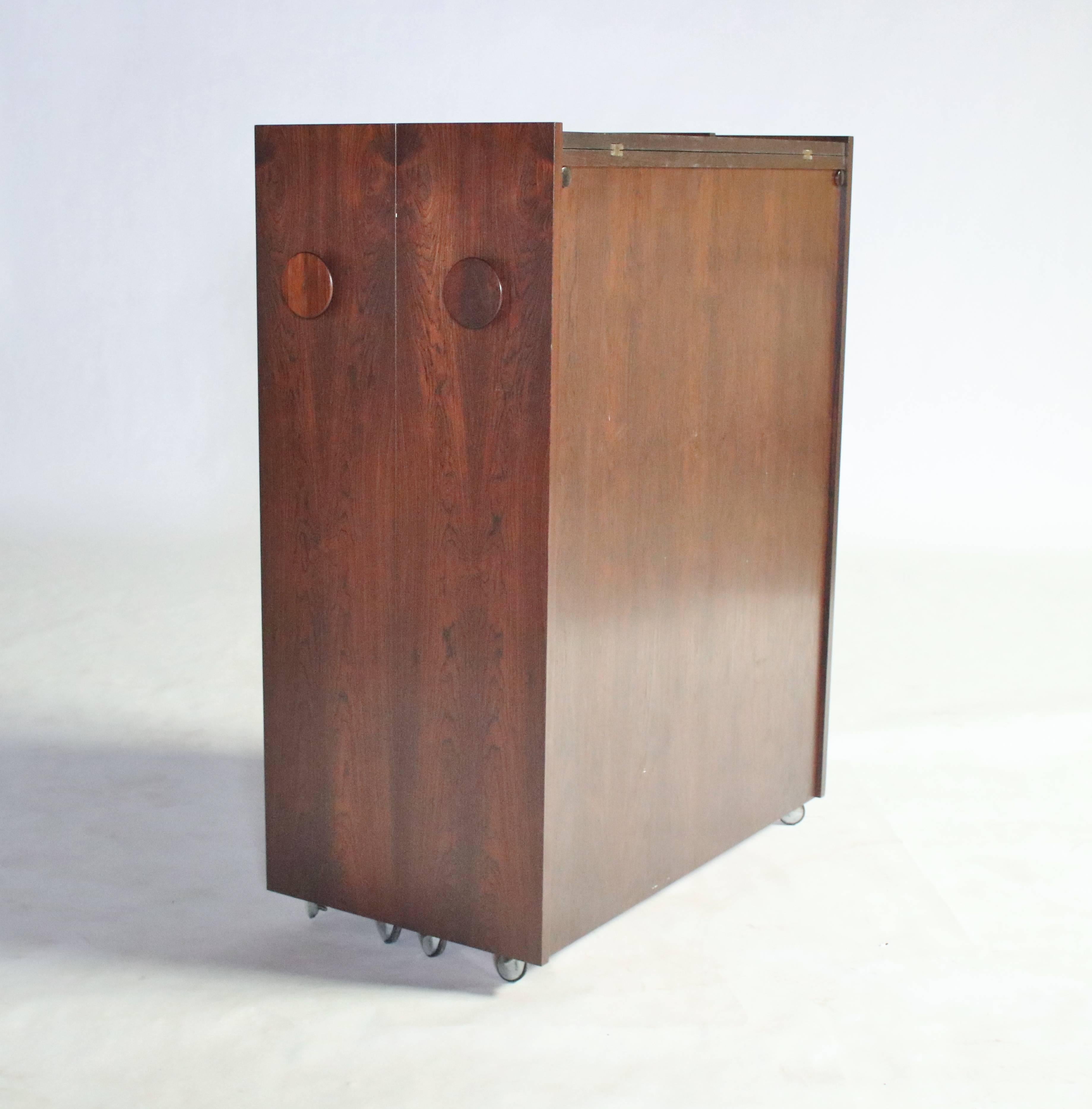 Folding Danish modern dry bar designed by Eric Buch in a beautiful rosewood case. Opens to a rosewood interior with three adjustable shelves and hinged over-hang shelves lined in black laminate for beverages. Another interior shelf open to reveal an