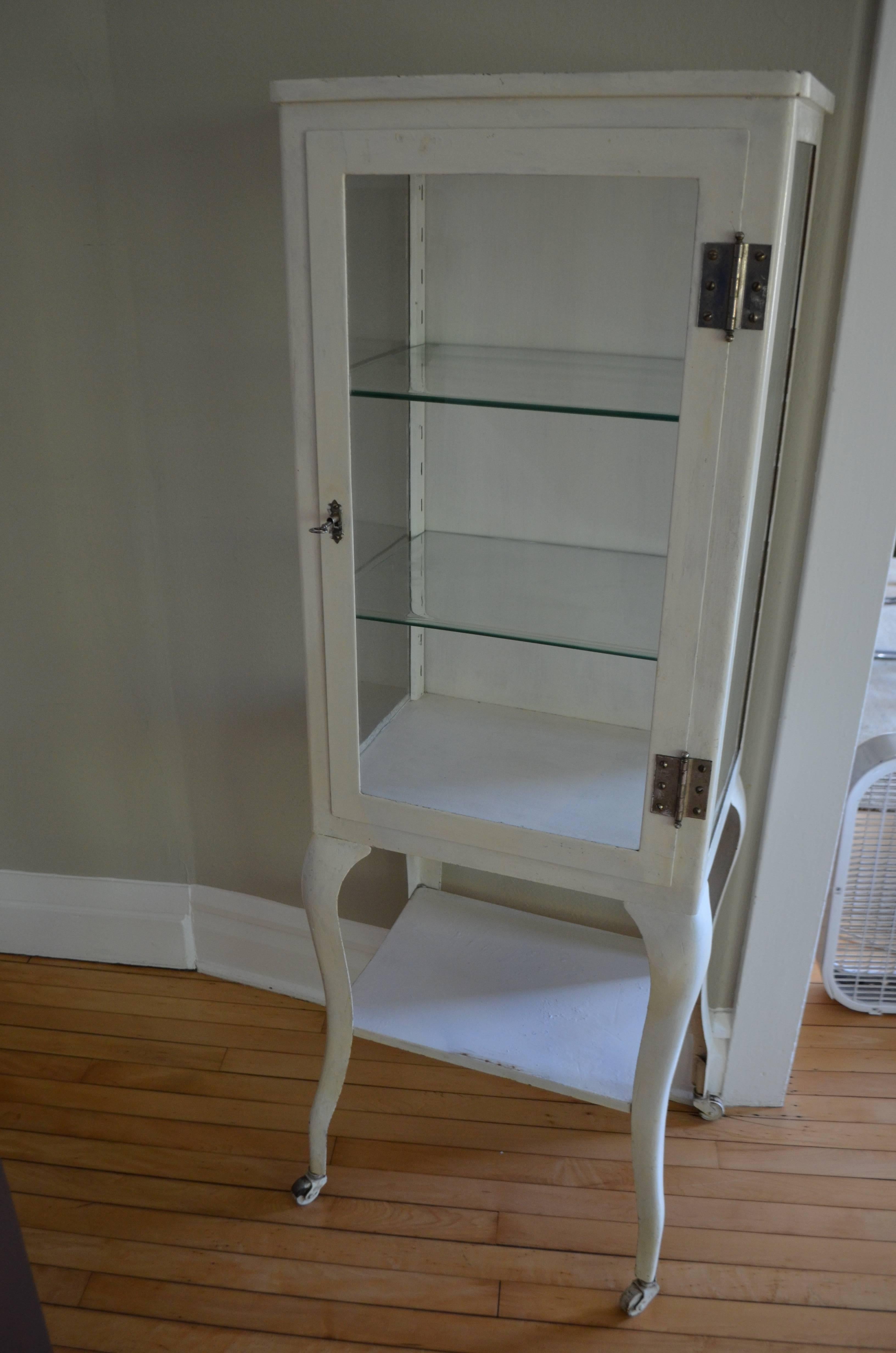 In as-found white enamel and with wheels. Original key included and works to lock the cabinet tightly. Two adjustable, original thick glass shelves with beveled edges. Use for storage or as bar. If you prefer, it could be stripped to bare steel and