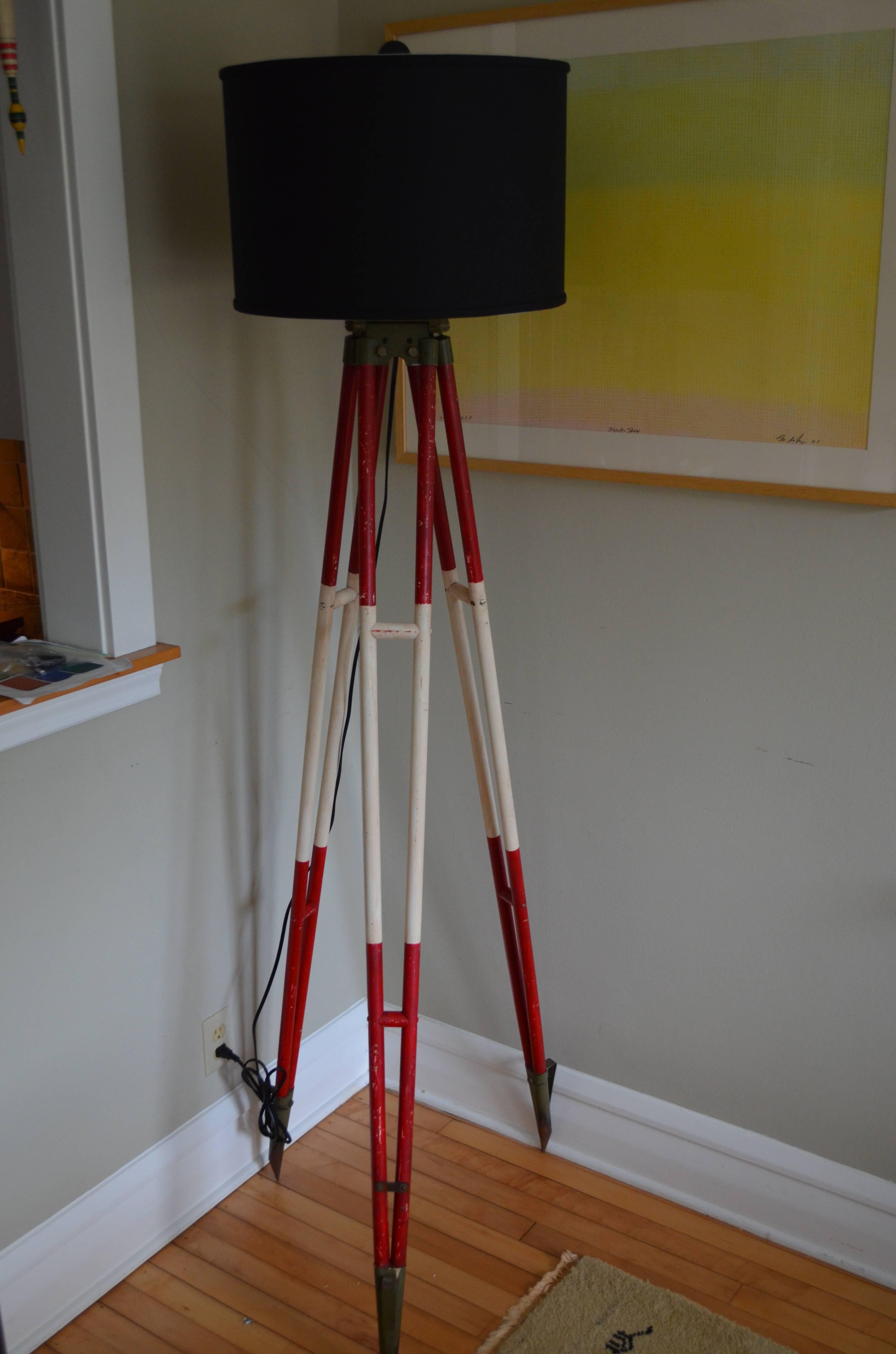 Floor lamp from Swiss surveyor tripod stands statuesque on steel-tipped feet and wooden legs in as-found red and white paint that stands out in the field. Professionally wired with UL-approved components including 10-foot black cord, three-way