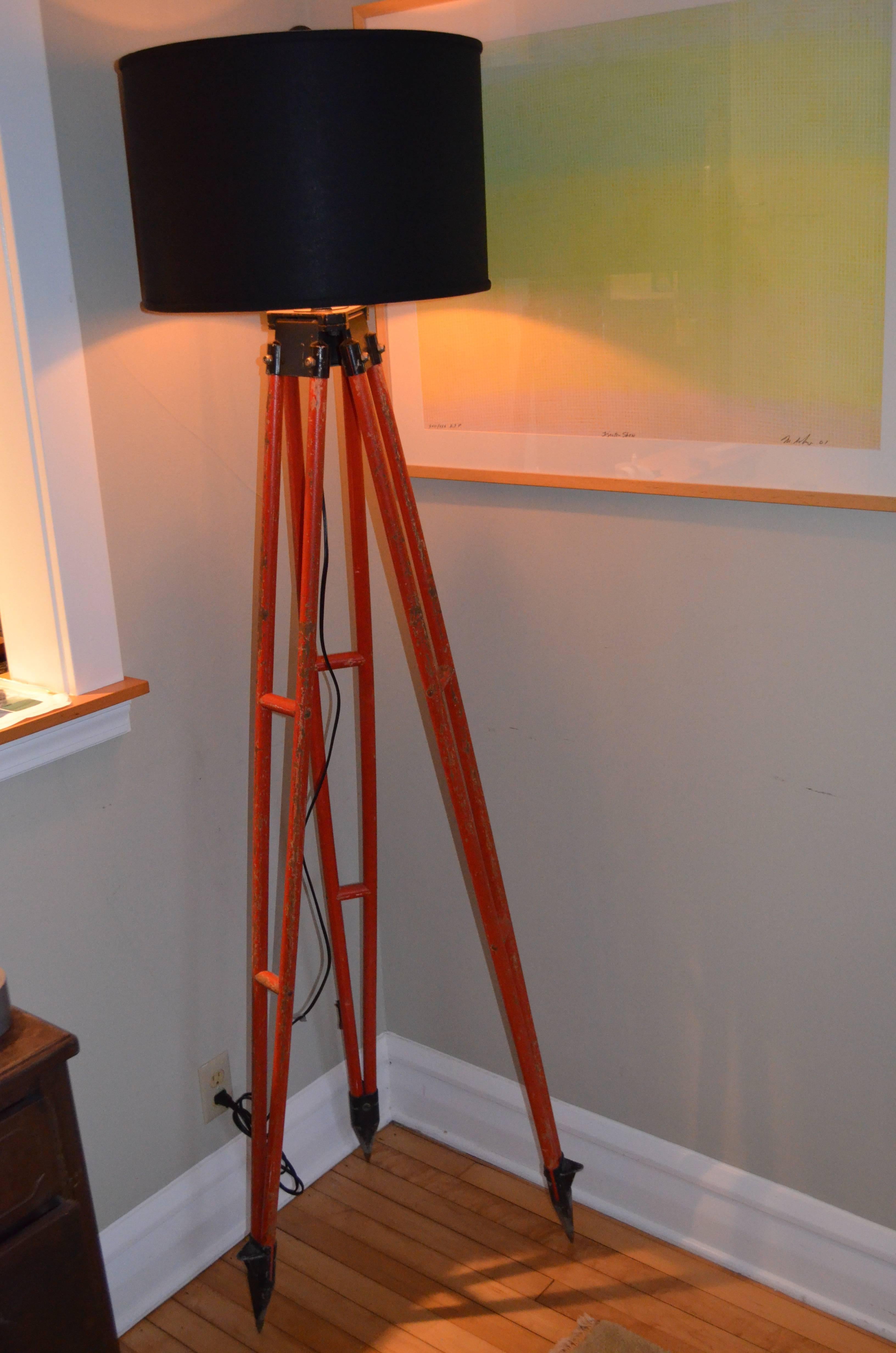 Floor lamp from surveyor's tripod stands statuesque on steel-tipped feet and wooden legs in as-found orange paint that stands out in the field. Professionally wired with UL-approved components including ten-foot black cord, three-way switch socket