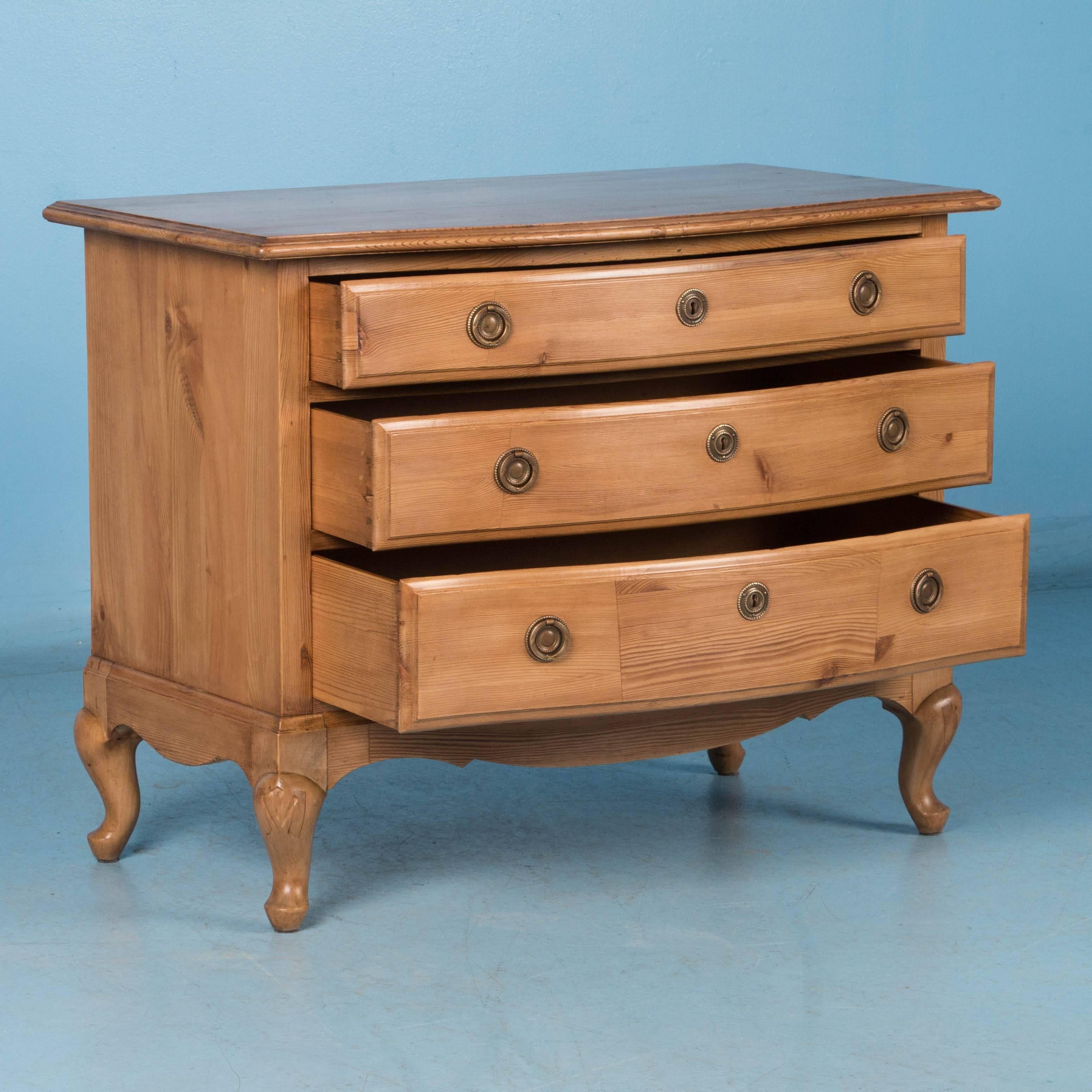 Small with lots of charm, this chest of drawers in natural pine has been given a waxed finish, bringing out the warmth of the wood. It features a slightly bowed front with three graduated drawers, brass hardware and carved cabriole legs. The case is