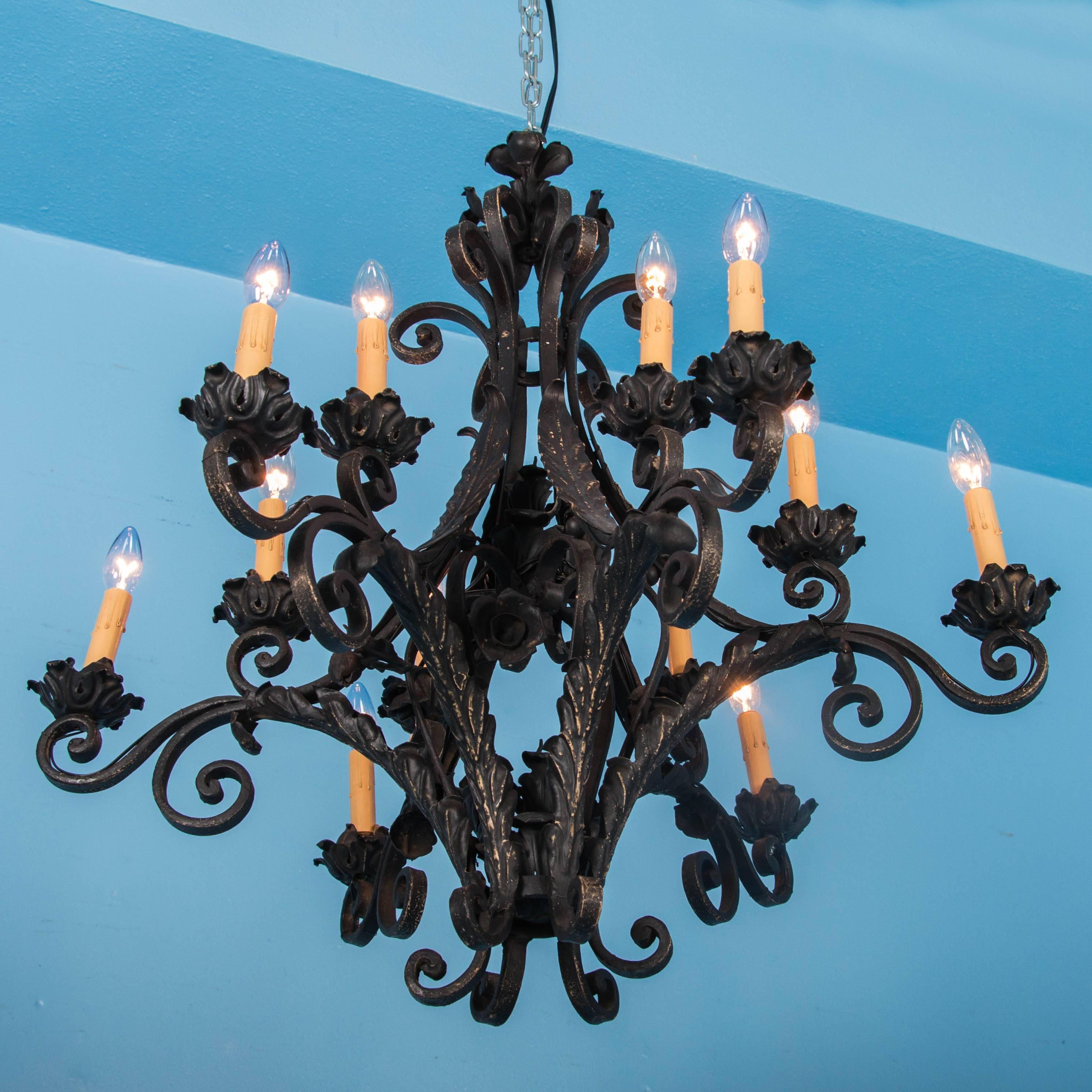 This antique French wrought iron chandelier, circa 1920, has six arms that radiate out from the centre. Each arm is set with a pair of lights, all with a candle cover and resting on a floral drip pan. With scrolled arms and applied leaf details, the