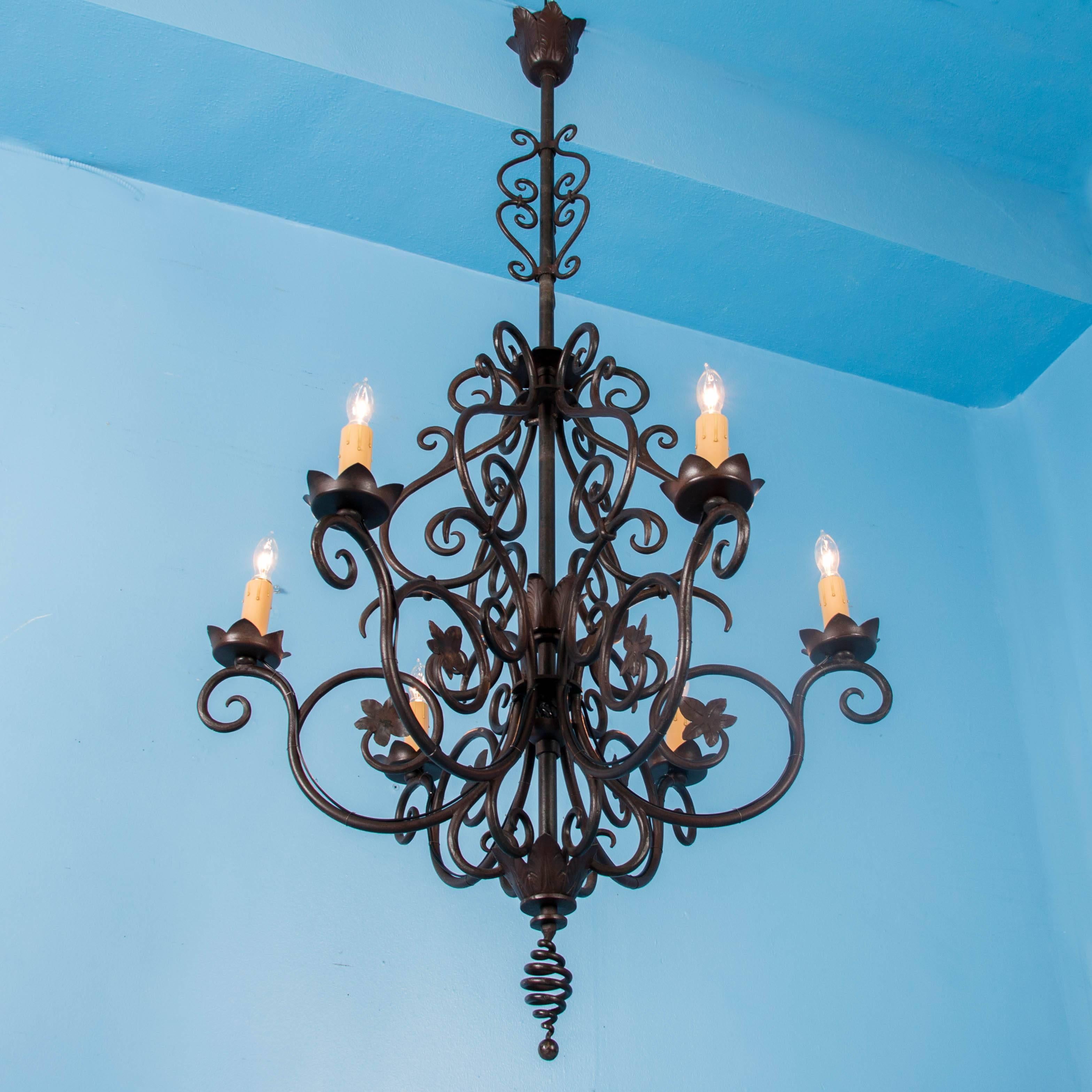 This large antique wrought iron chandelier features a long central rod supporting the six ornate scrolled arms and applied leaf details. Each of the six sockets are covered with a candle cover which rests on a round leaf drip pan. The chandelier has