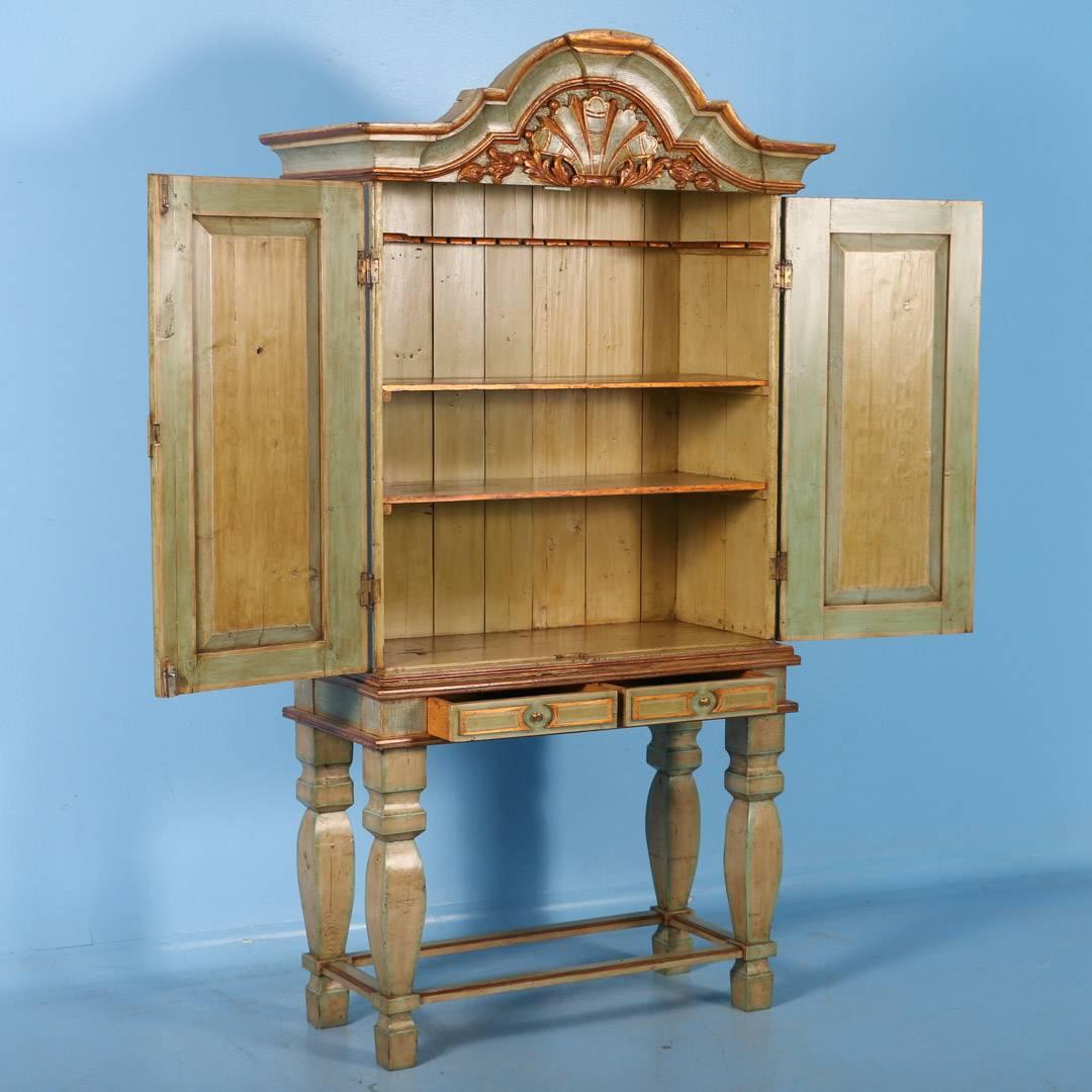 This Baroque Cabinet is crowned by a dramatic curved top with carved foliage. The cabinet dates between 1820-40, while the paint was added later. The soft green has blue undertones while bits of red appear under the gold gilt. Please examine the