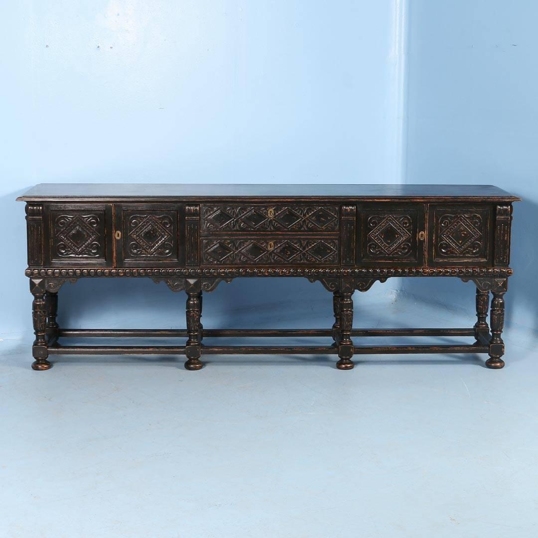 This dramatic long oak sideboard or buffet has carved panels, doors, drawers and even legs. It has recently been given a black painted finish and slightly distressed at edges, making the carvings and overall appearance even more striking. It has two